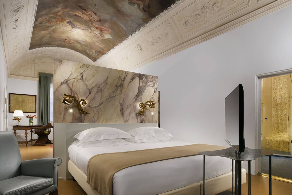 A hotel room with a large bed, a television, a marble wall, and a ceiling with frescoes.
