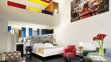 Una habitación de hotel moderna con una cama grande, un sillón rojo, una mesa de centro de cristal y un cuadro en la pared.