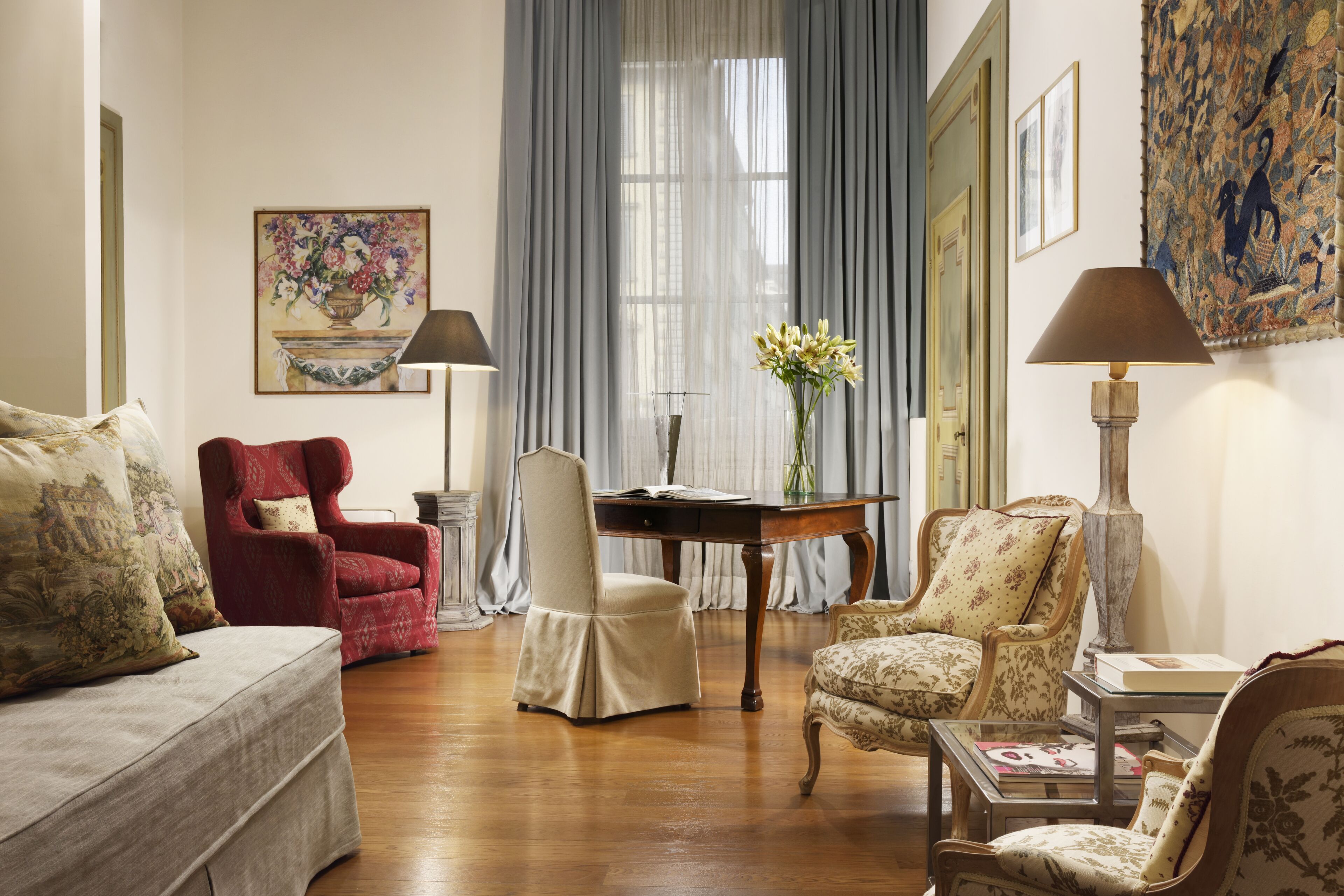 A traditional room with a wooden floor, a red armchair, a white chair, a beige sofa, and a wooden table with a vase of flowers.