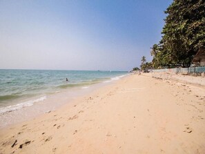 Uma praia arenosa com pegadas que levam à água, céu claro e algumas pessoas à distância.