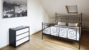 A bedroom with a black metal bed frame, a white bedside table with drawers, a black and white cityscape painting on the wall, and a skylight above the bed. - Hotel Am Lindenhof (Hannover)
