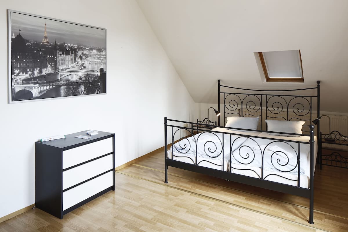 A bedroom with a black metal bed frame, a white bedside table with drawers, a black and white cityscape painting on the wall, and a skylight above the bed.
