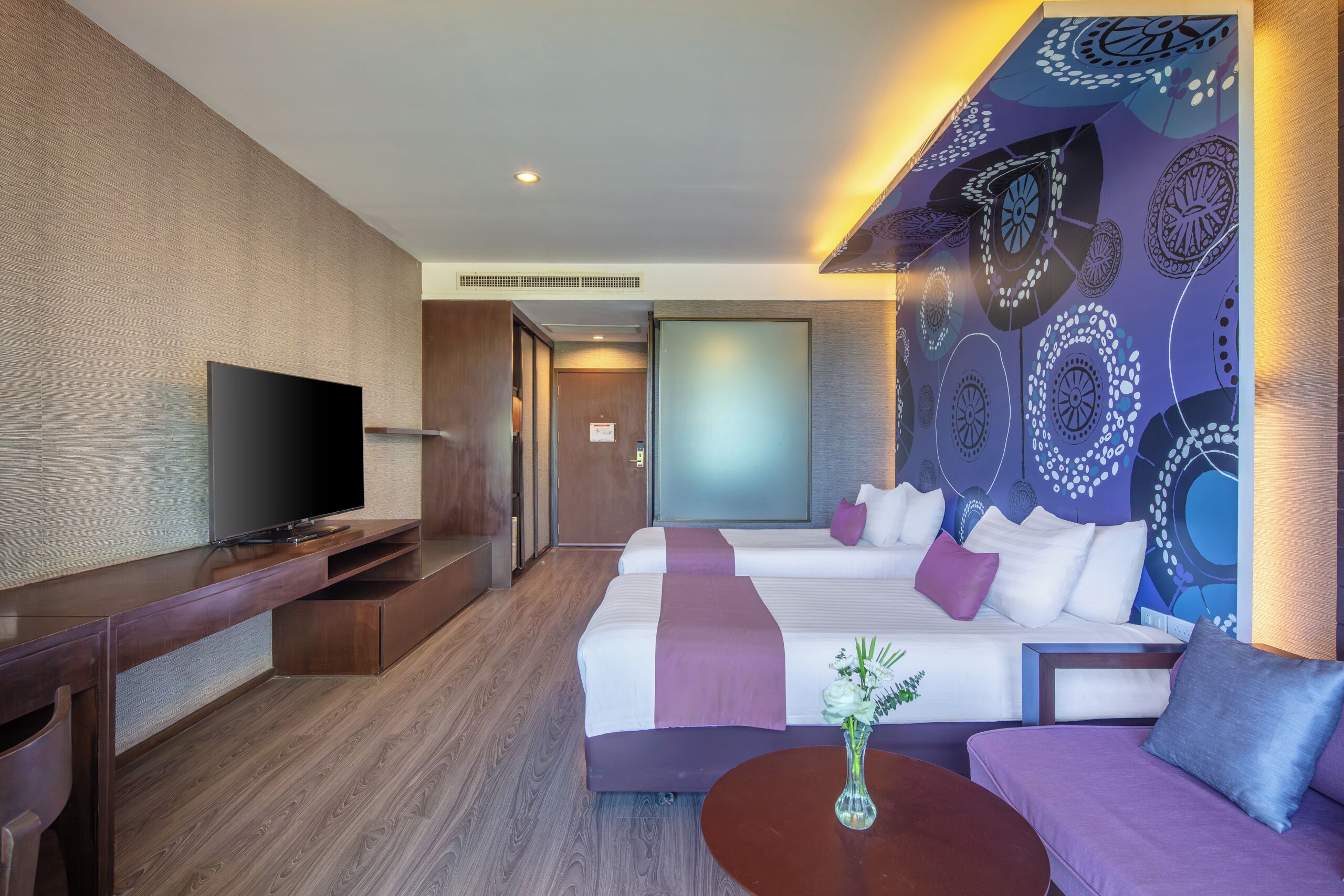 A hotel room with a flat-screen TV, two beds with purple and white bedding, a coffee table with a vase, and a patterned wall.