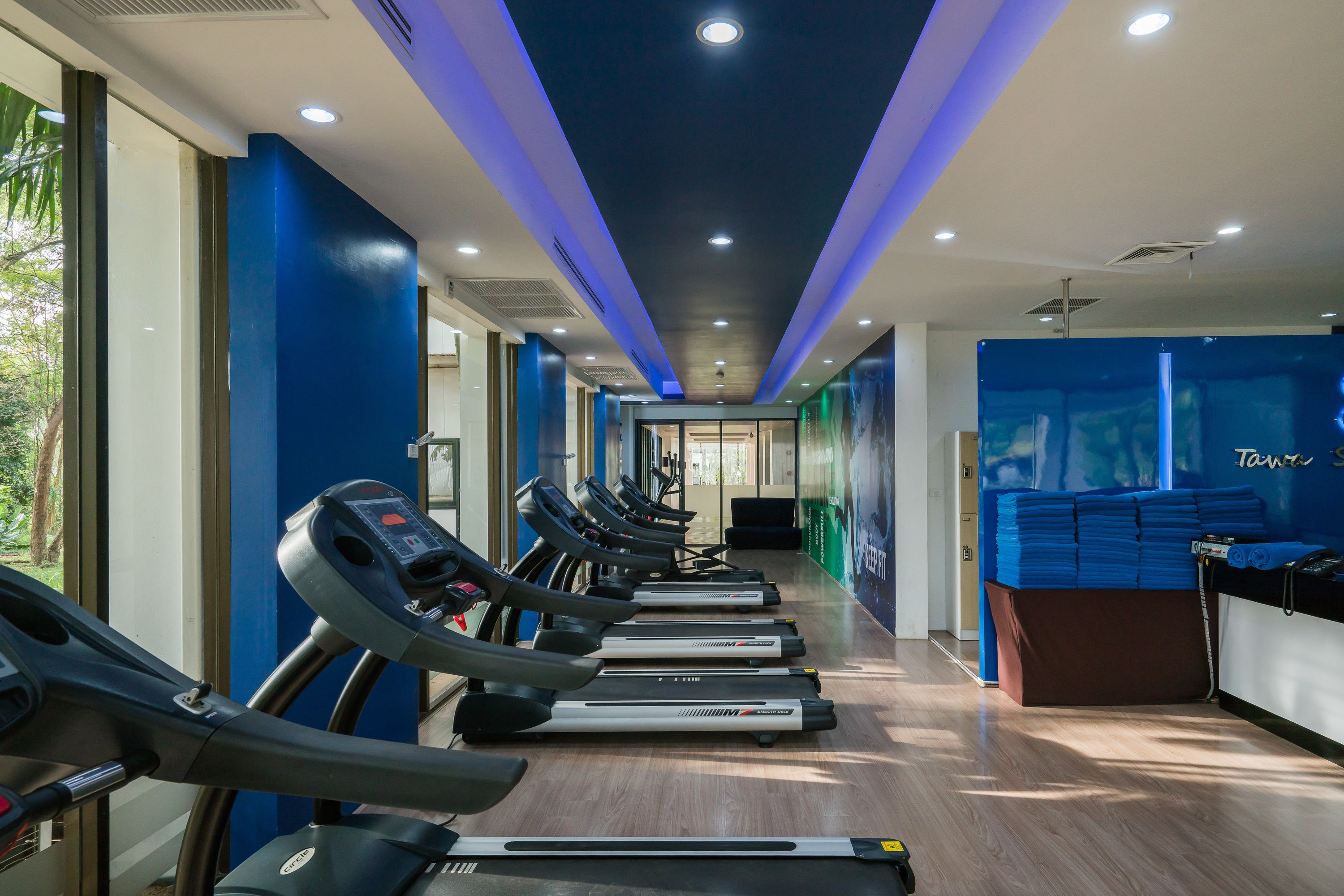 Une salle de sport avec des tapis de course alignés le long d’un couloir, un comptoir d’accueil et un mur bleu orné d’une affiche.