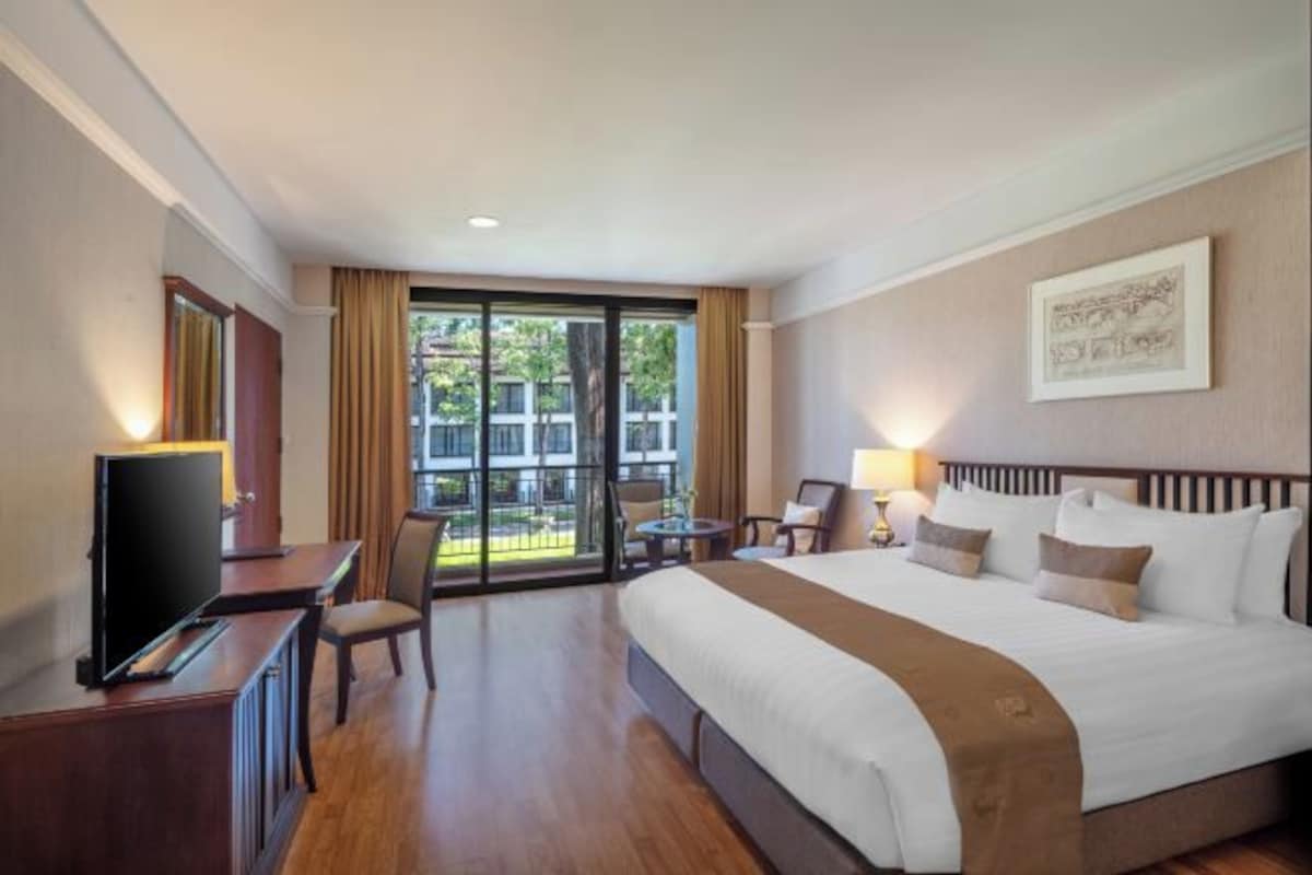 A hotel room with a large bed, a desk with a television, a chair, a small table, and a balcony with a view of buildings.