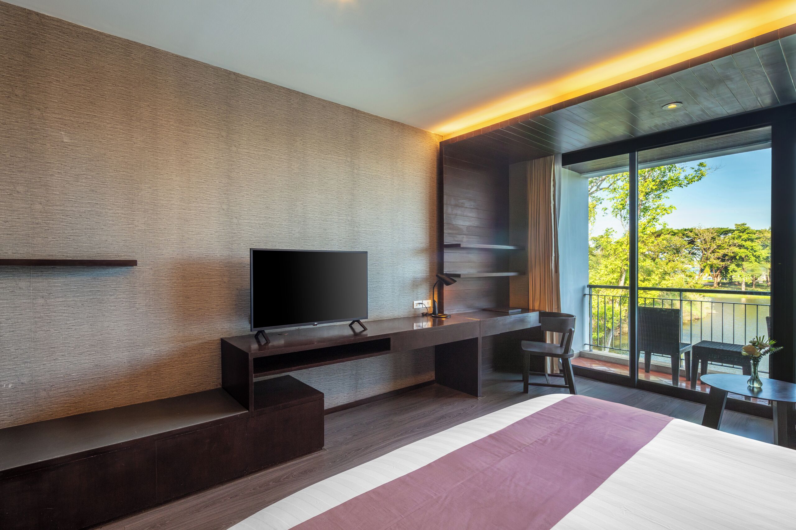 A modern hotel room with a large bed, a flat-screen TV, a desk with a chair, and a balcony with a view of trees.