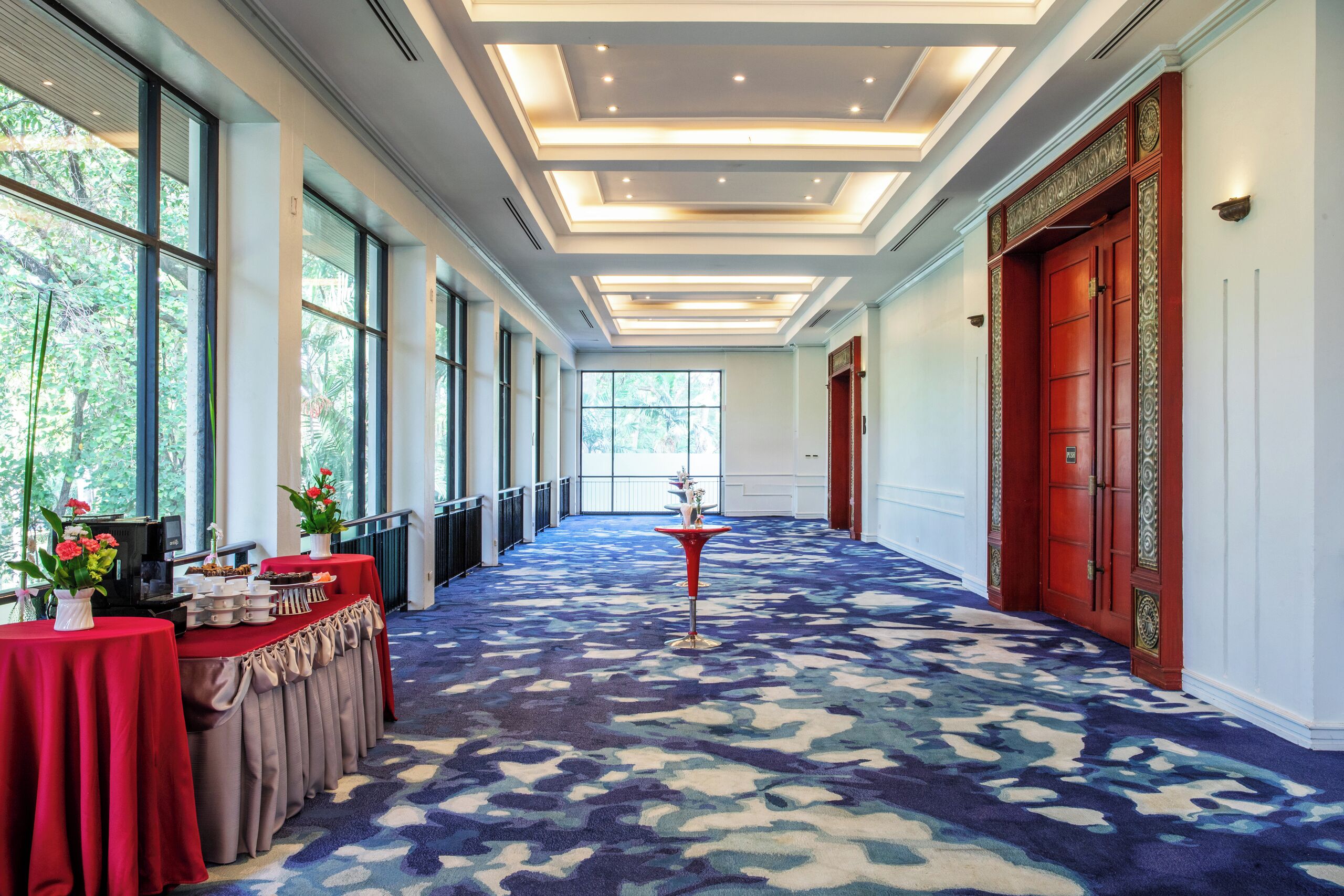 A hotel corridor with a patterned carpet, large windows, and a red door.