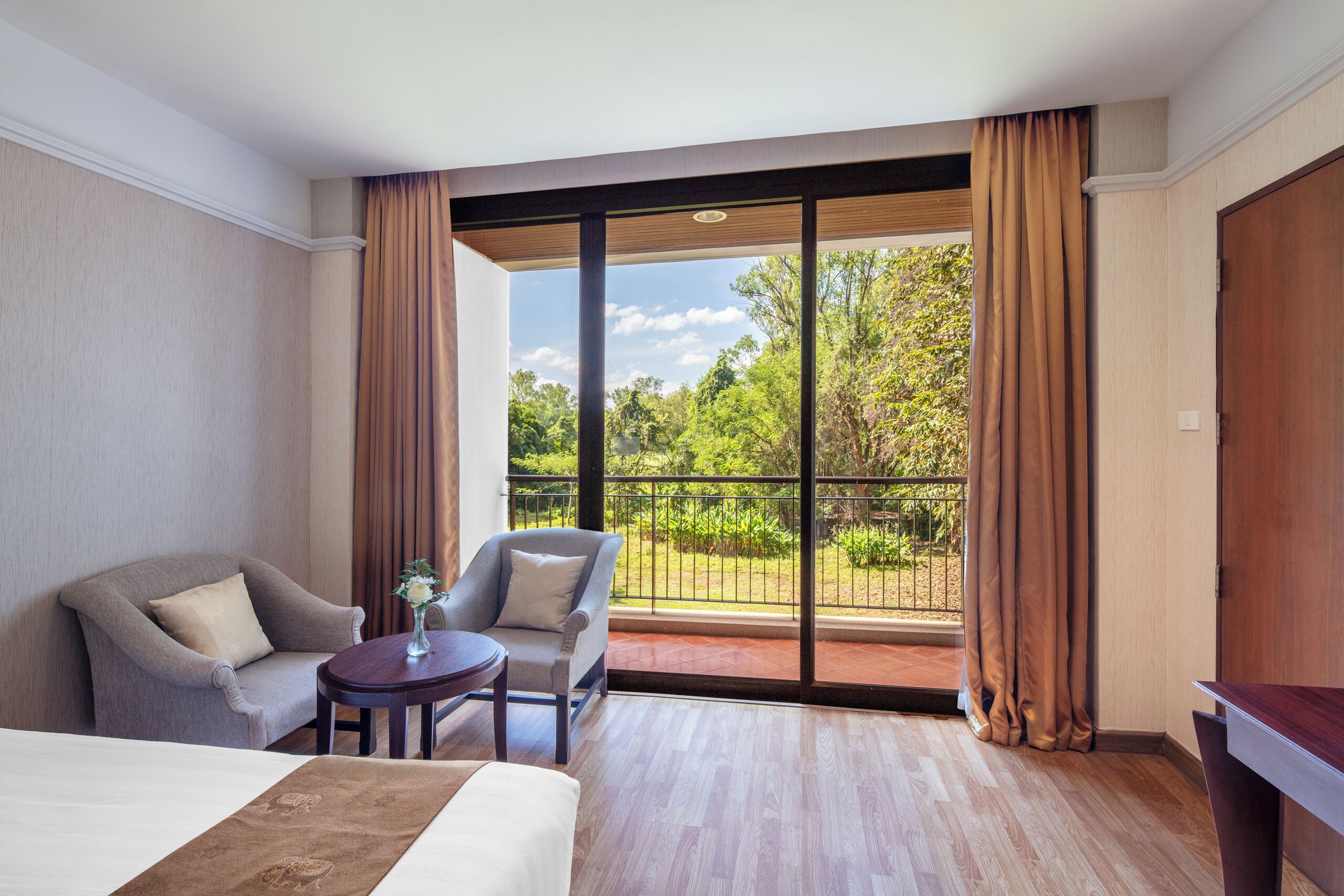 A hotel room with a bed, two armchairs, a small table, and a view of the outdoors through large windows.