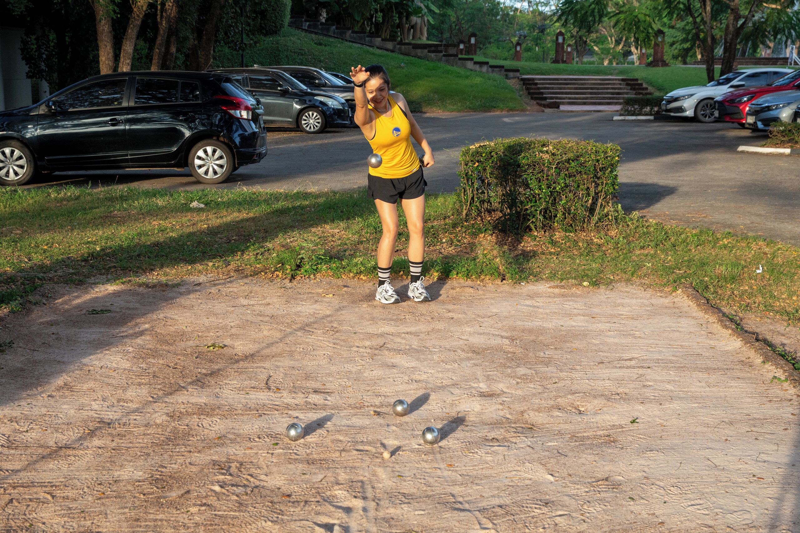 A person playing with balls on a concrete path near parked cars and greenery.