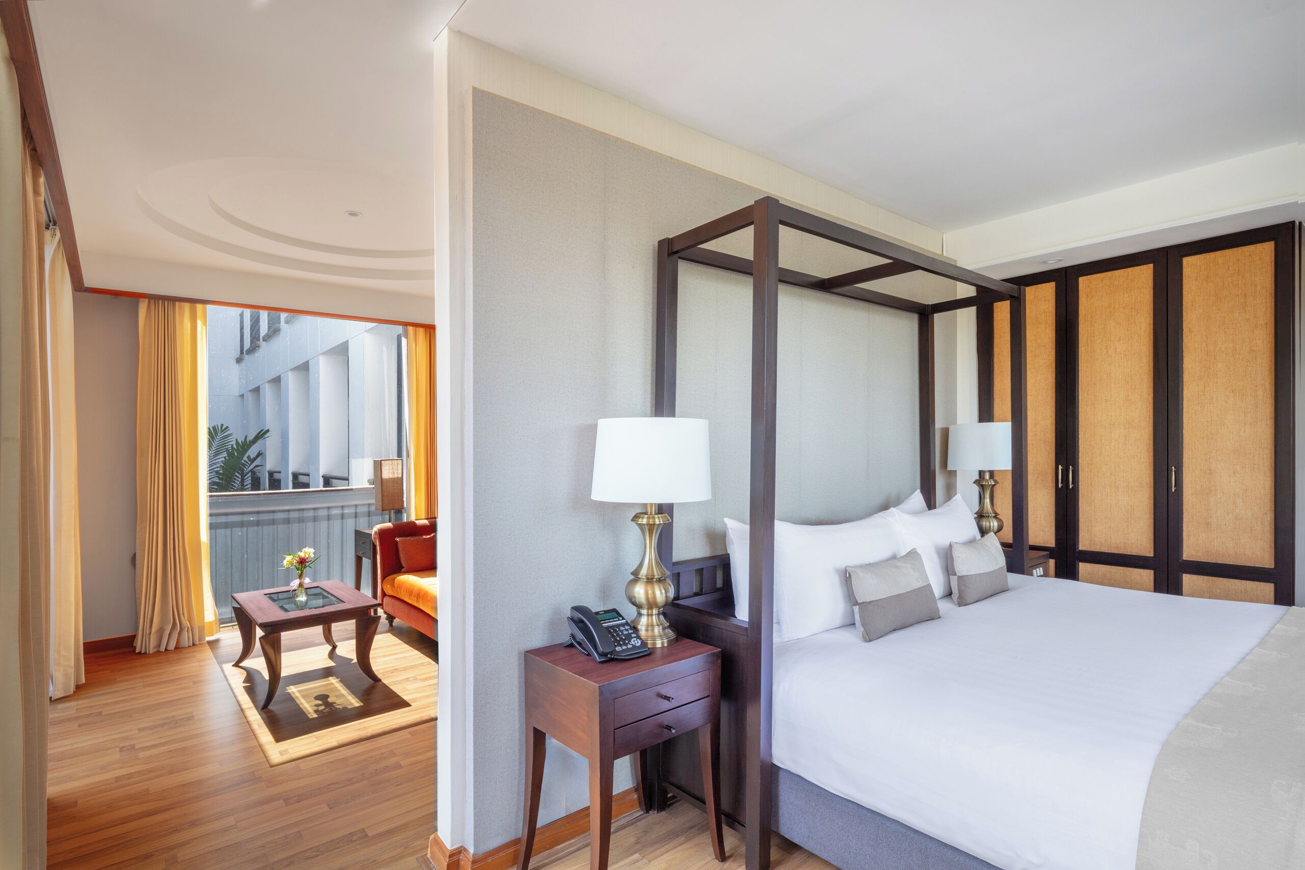A hotel room with a large bed, a bedside table with a lamp, a small table with a phone, and a view of the outside balcony.