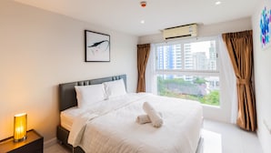 A hotel room with a bed, a nightstand, an air conditioning unit, a window with curtains, and a city view. - The Place Pratumnak by PSR ASIA (Pattaya)