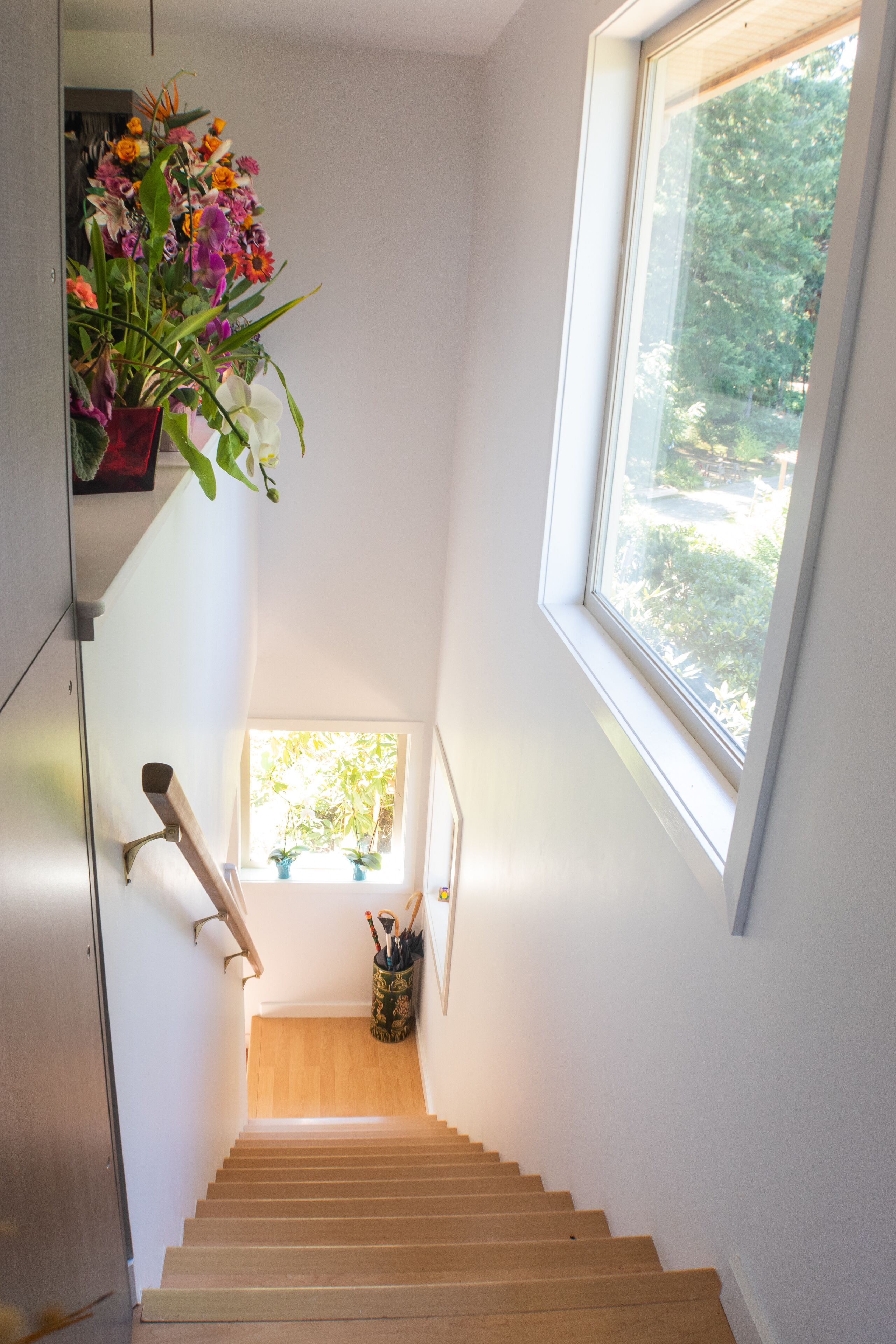 A staircase with a wooden handrail, a window with a view of greenery, and a hanging flower arrangement.