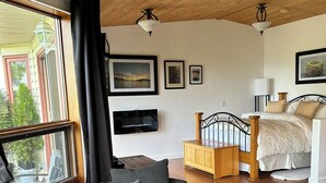 A cozy room with a wooden ceiling, a large window with curtains, a grey armchair, a bed with a wooden headboard, and a small wooden cabinet. - Elevate Rooms Armand Heights (Salt Spring Island)