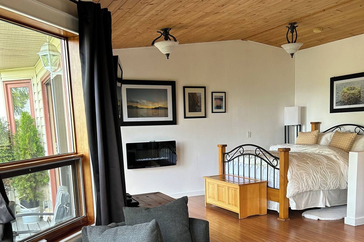 A cozy room with a wooden ceiling, a large window with curtains, a grey armchair, a bed with a wooden headboard, and a small wooden cabinet.