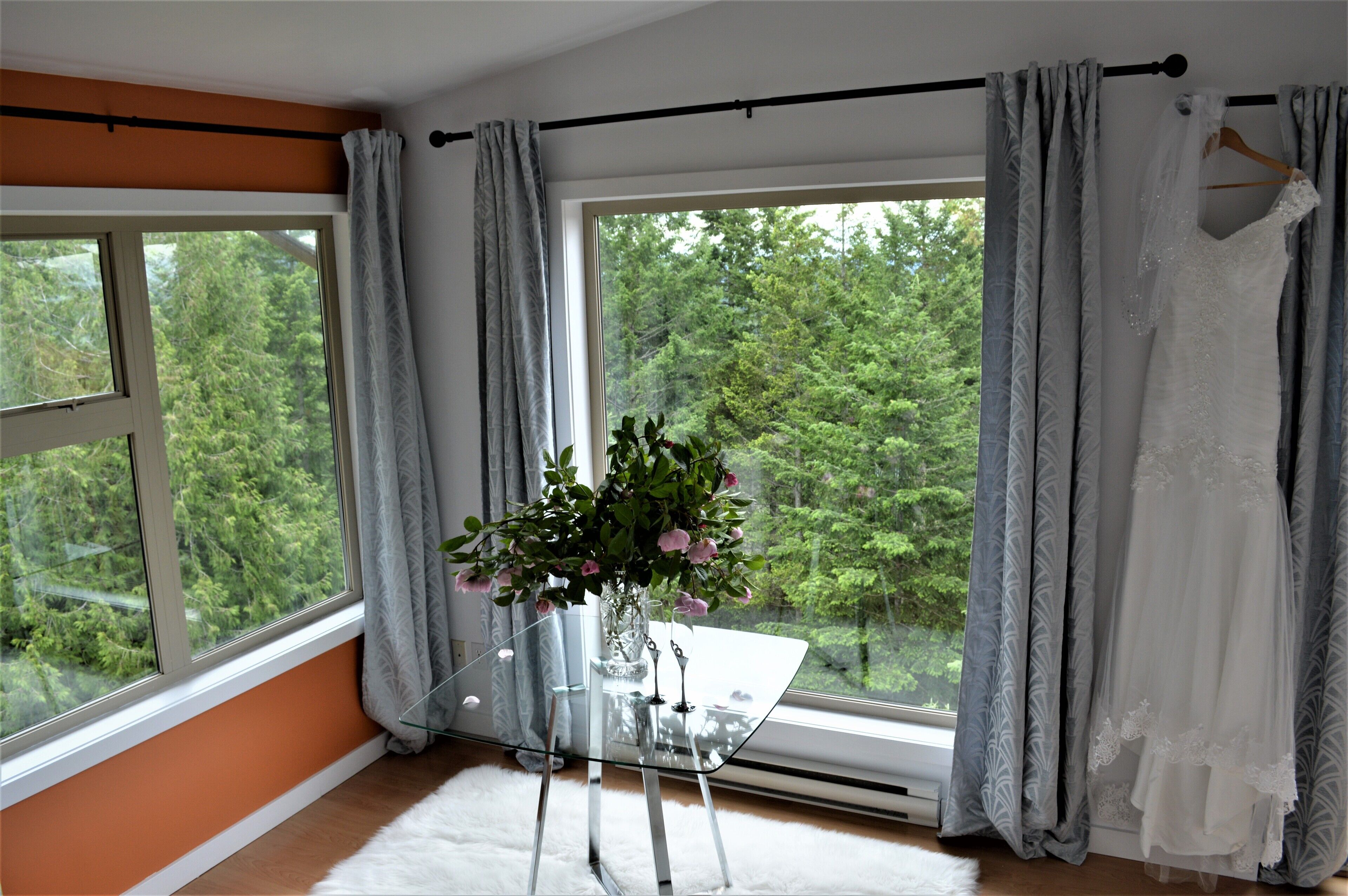 A room with a large window, a glass table with a vase of flowers, and a wedding dress hanging on a curtain rod.