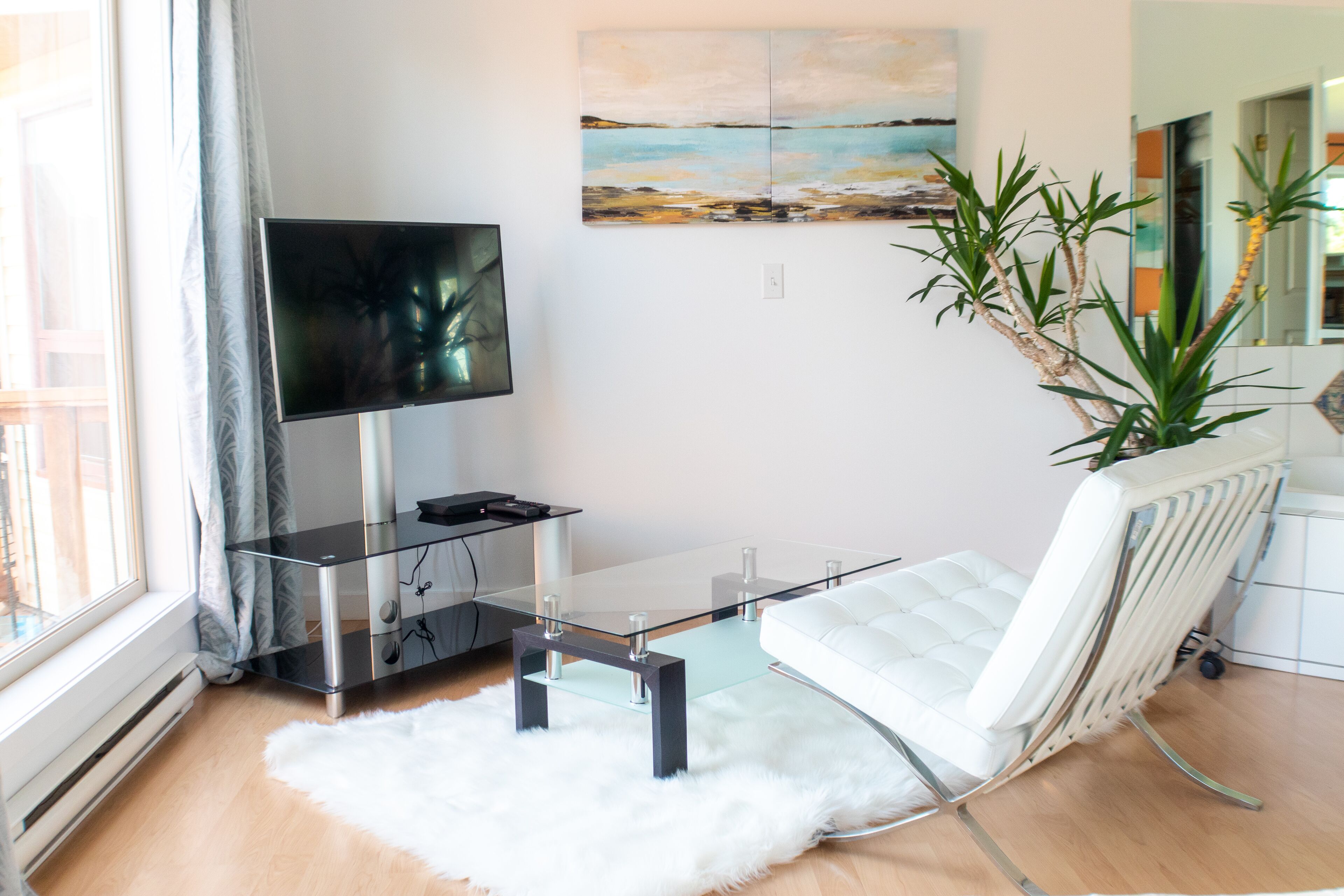 A modern living room with a glass coffee table, a white sofa, a flat-screen TV, a painting on the wall, and a potted plant.