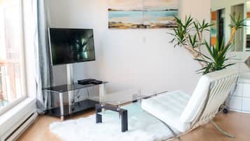 A modern living room with a glass coffee table, a white sofa, a flat-screen TV, a painting on the wall, and a potted plant.