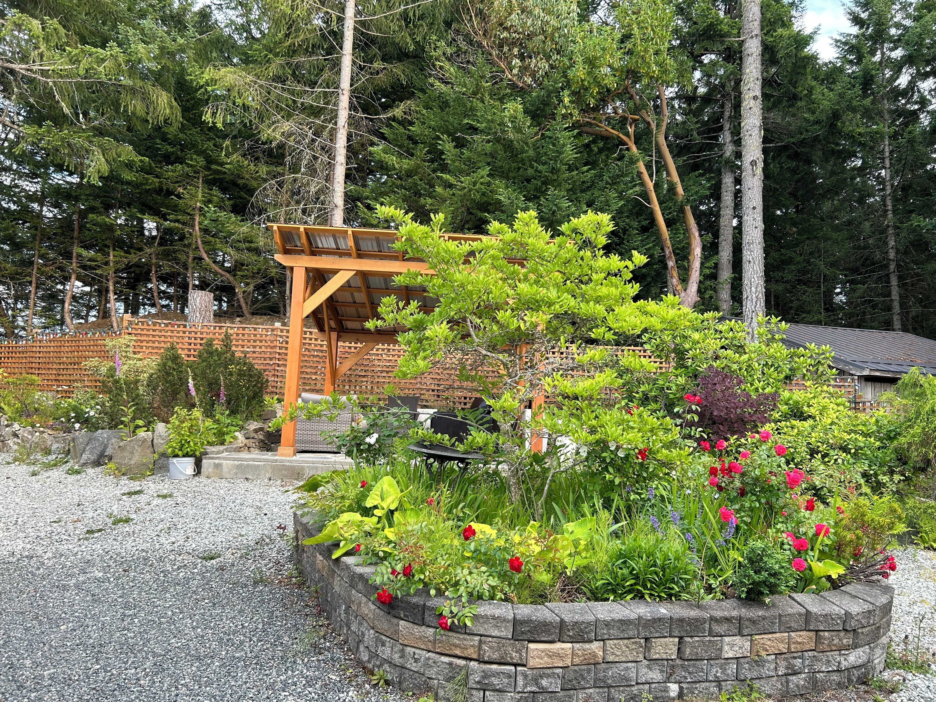 Un jardín con un porche de madera, un muro de contención de piedra y diversas plantas y flores.