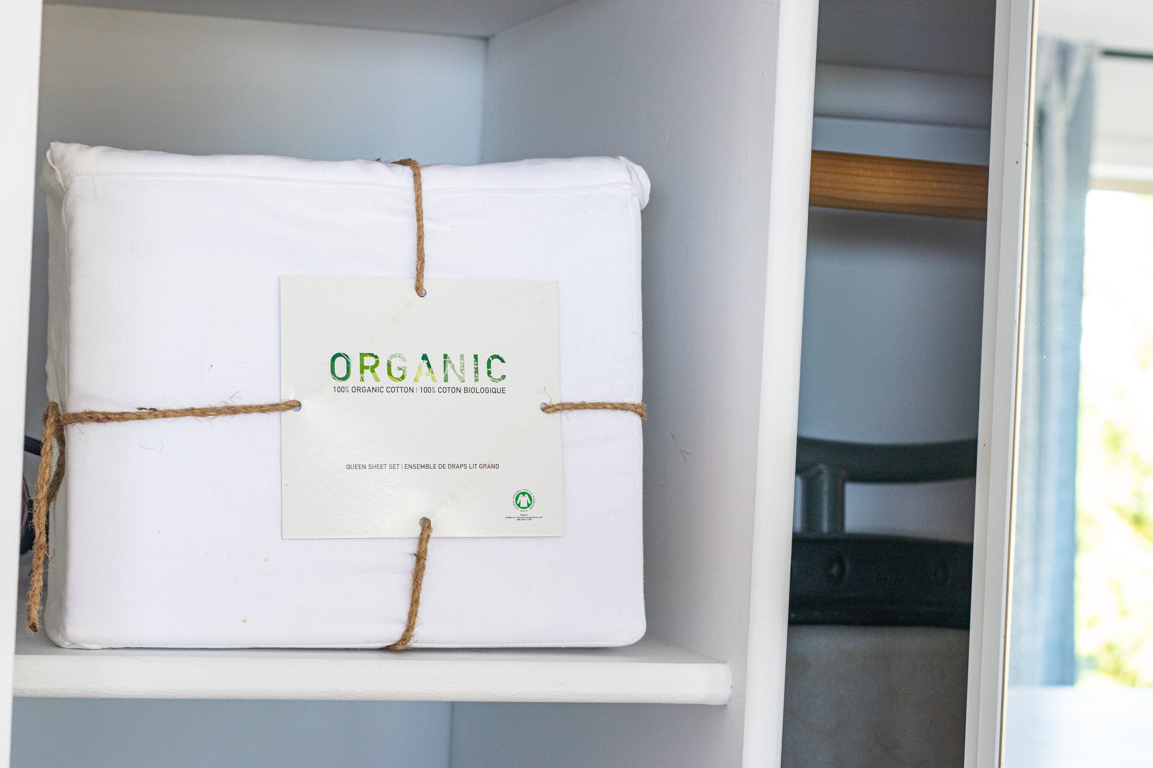 A white shelf with a white package tied with twine, labeled "ORGANIC 100% ORGANIC COTTON 100% COTON BIOLOGIQUE QUEEN SHEET ENSEMBLE DE DRAPS LIT GRAND."