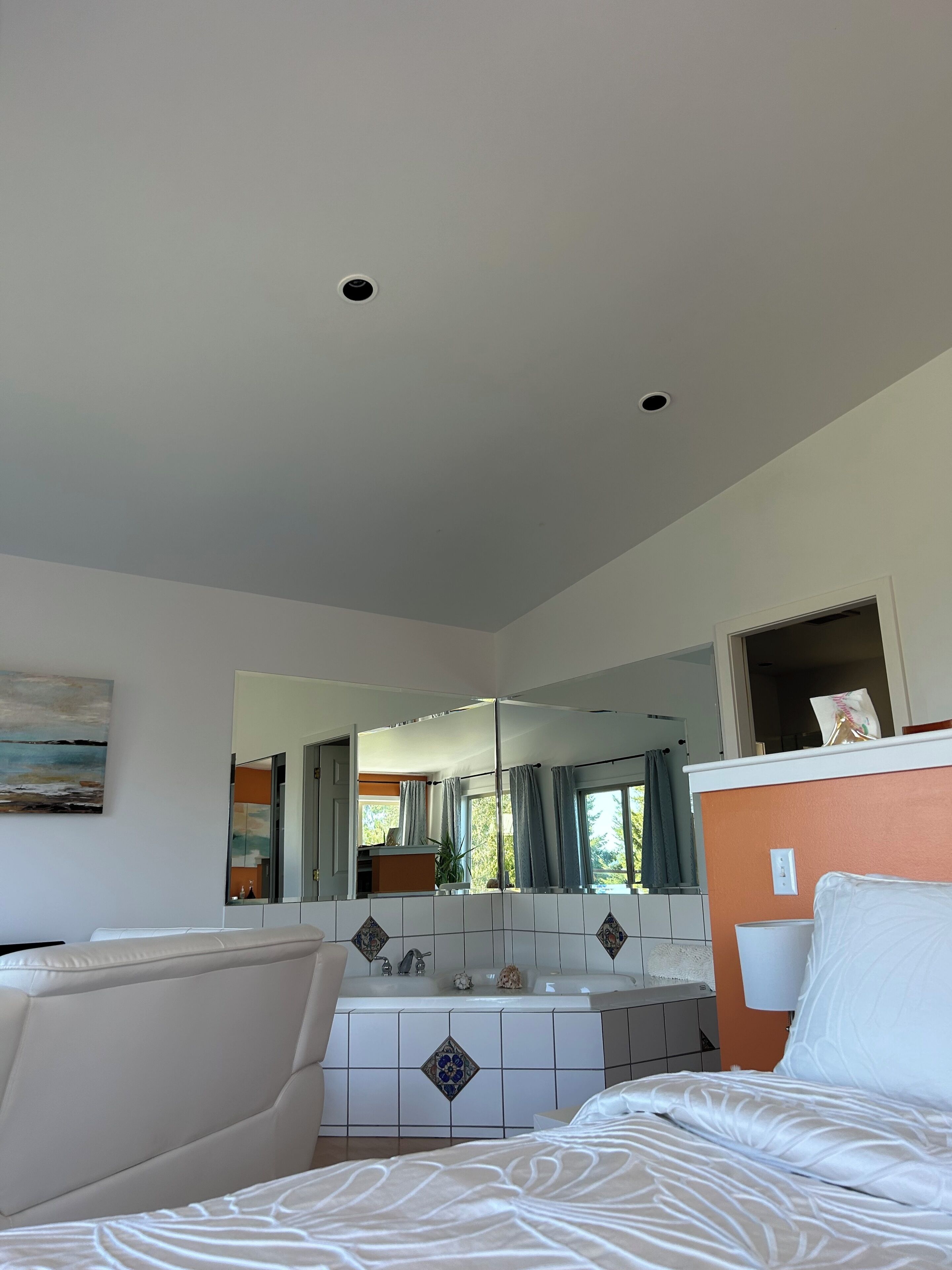 A modern bedroom with a large bed, a white dresser, a bathroom with a sink and mirror, and a painting on the wall.