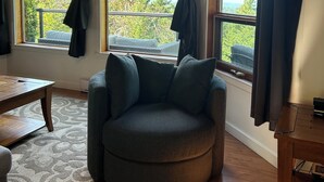 A cozy room with a wooden ceiling, a round grey armchair, a wooden table, and a view of greenery outside through large windows with dark curtains. - Elevate Rooms Armand Heights (Salt Spring Island)
