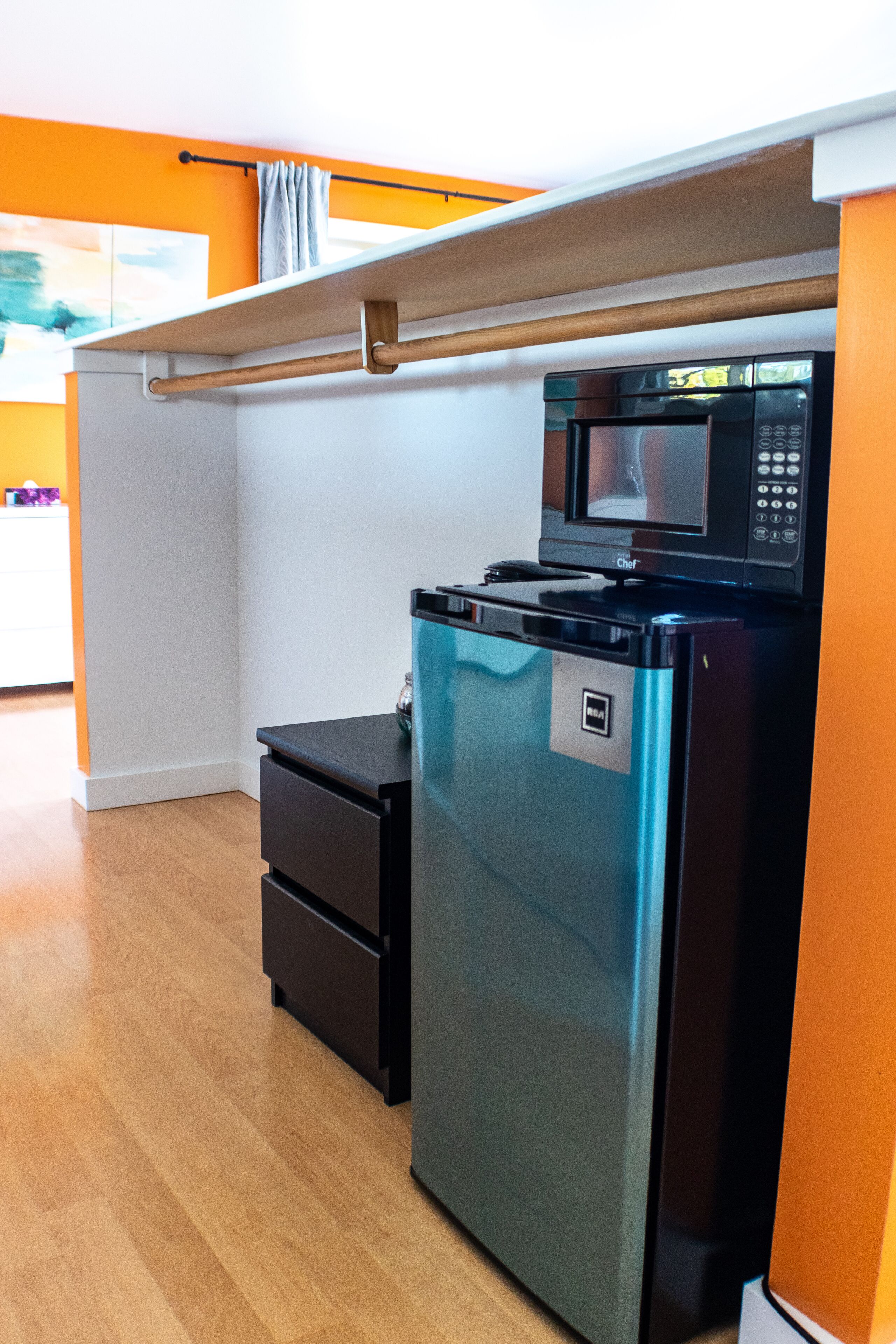 A compact kitchen with a built-in microwave and refrigerator, wooden flooring, and a white wall with a window.