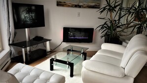 A modern living room with a glass coffee table, white leather sofa, and a flat-screen TV mounted on the wall. - Elevate Rooms Armand Heights (Salt Spring Island)