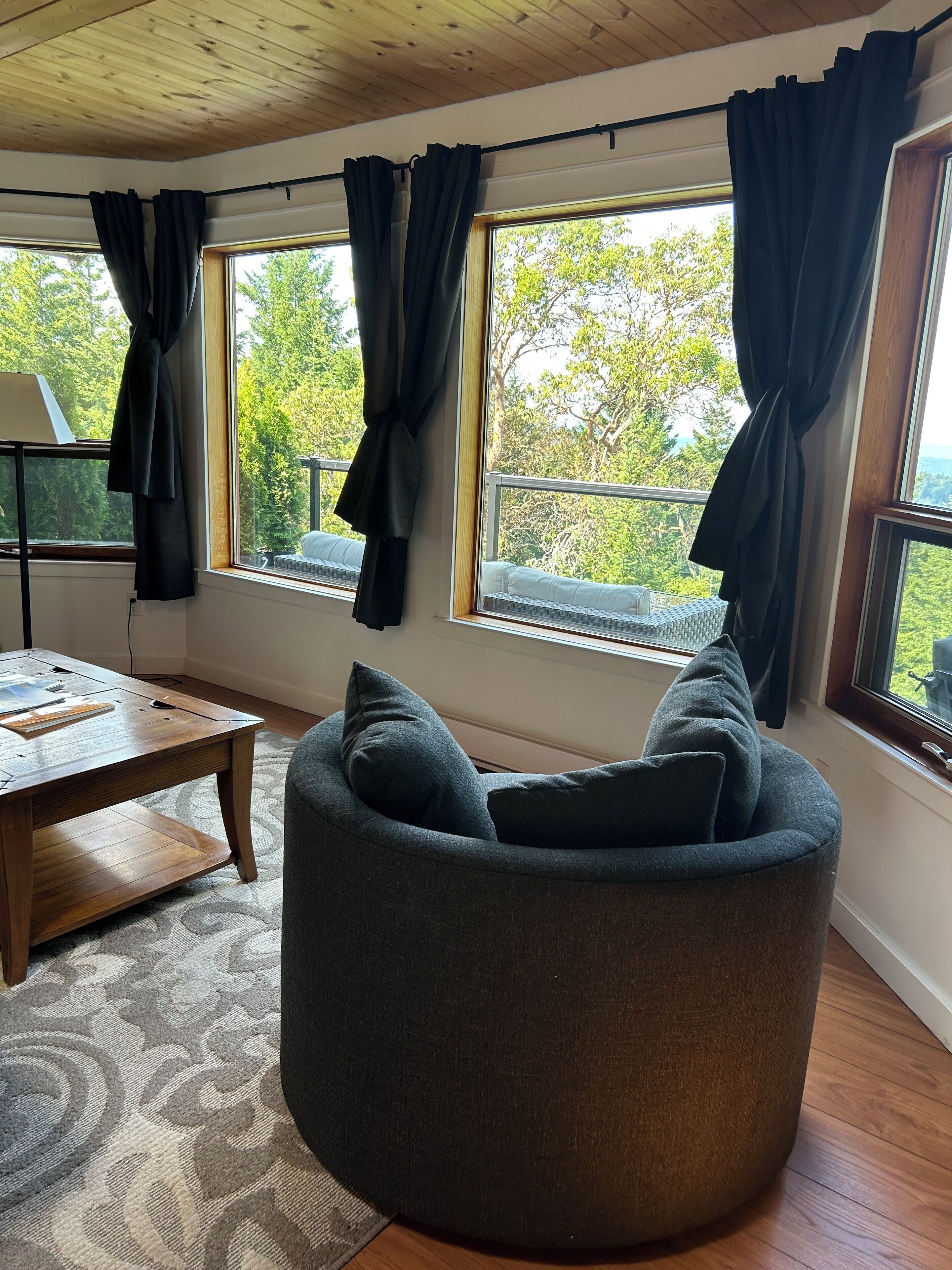 Un salón acogedor con un sofá redondo gris, una mesa de centro de madera y vistas a exuberante vegetación a través de amplias ventanas con cortinas negras.