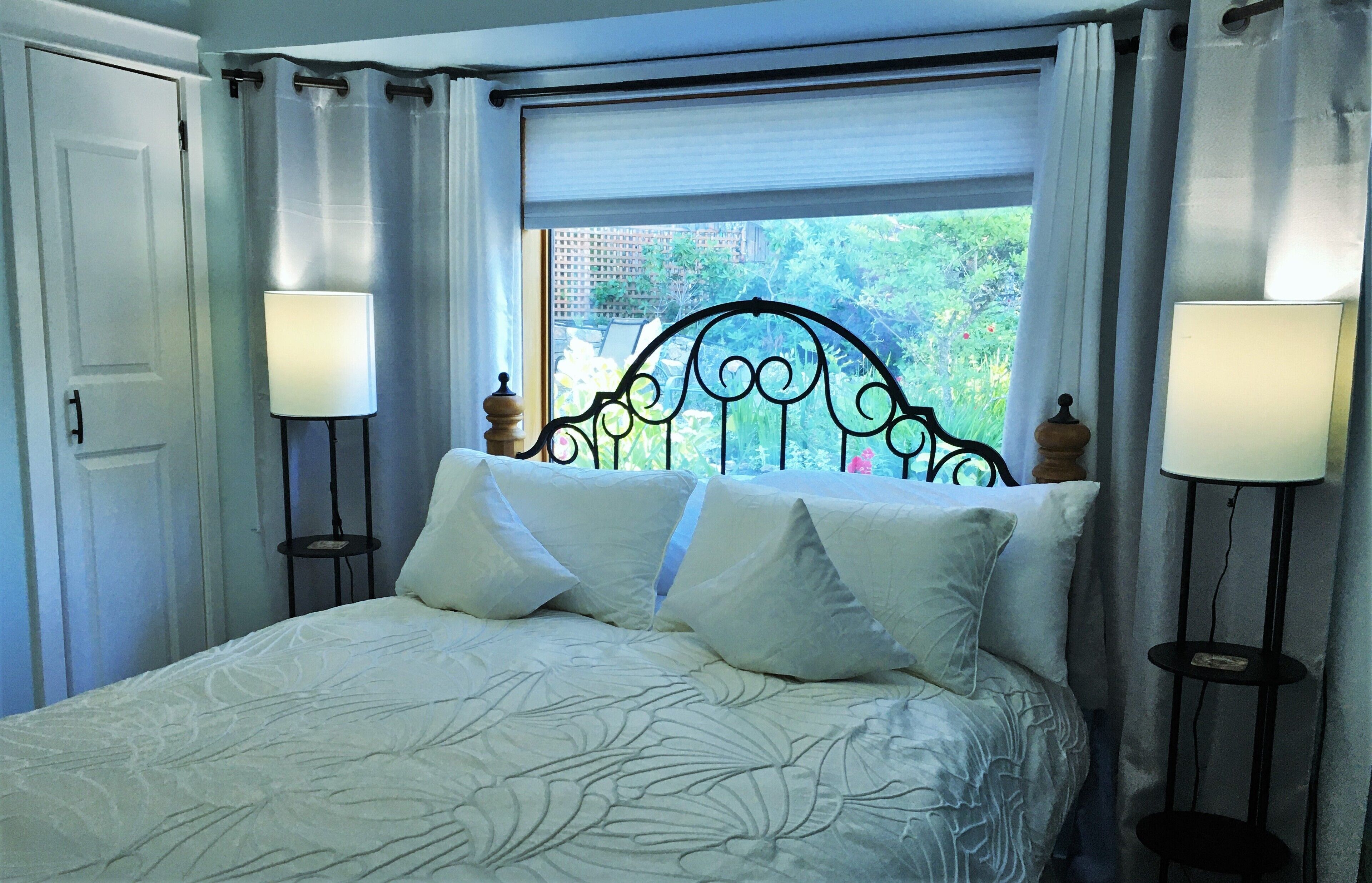 A neatly made bed with a patterned bedspread, two bedside lamps, and a window with a view of greenery outside.