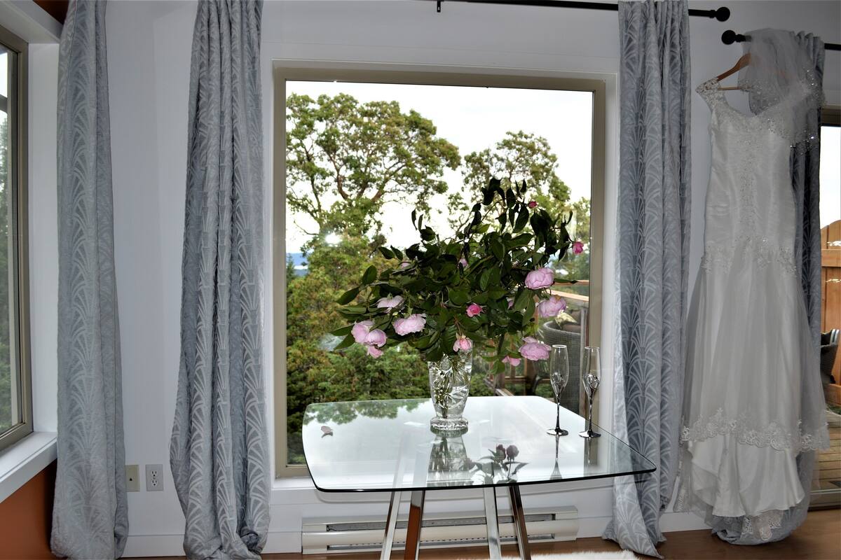 A room with a large window, a glass table with a vase of flowers, and a hanging garment.
