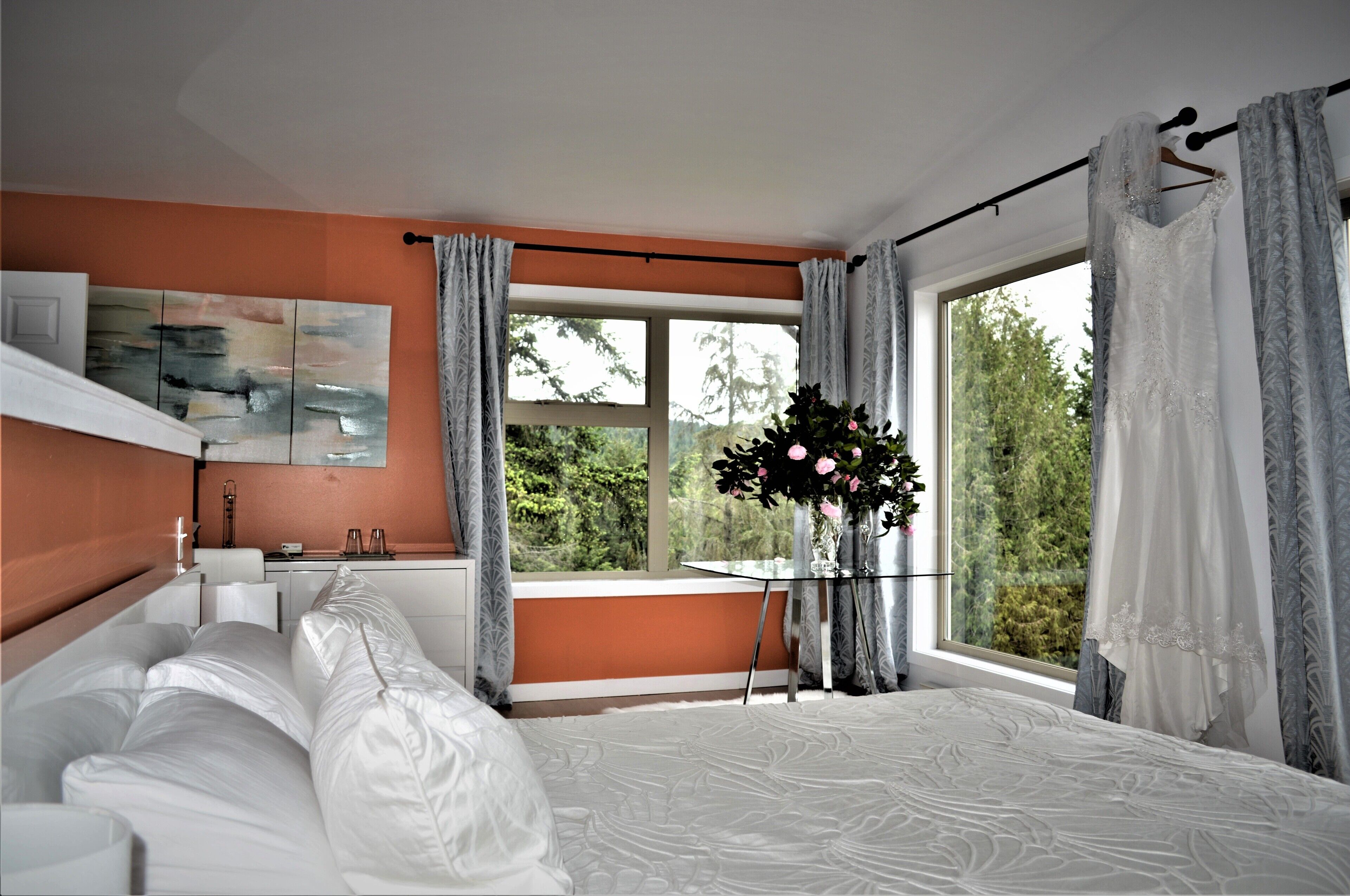 Un dormitorio con una cama grande, una ventana con cortinas, una mesita de noche con un florero y un cuadro en la pared.
