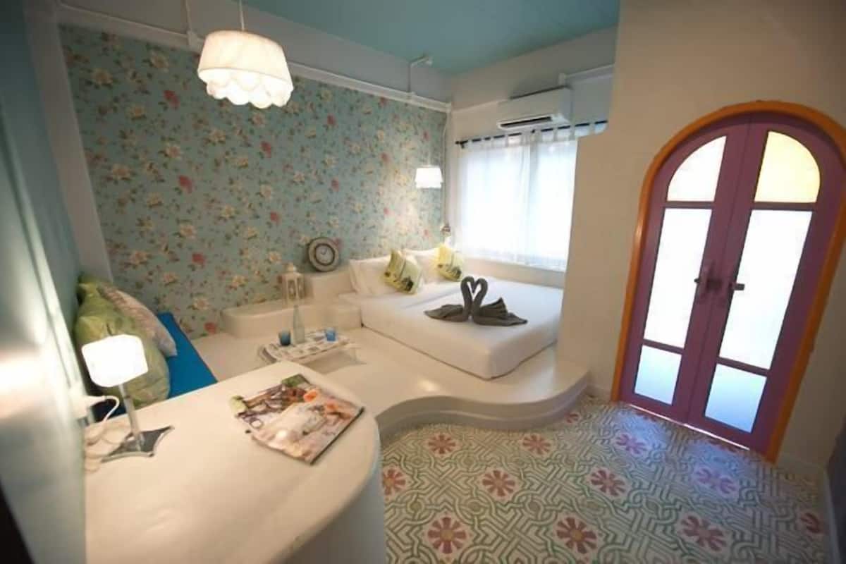 Um quarto com papel de parede floral, um sofá branco, uma mesinha de centro com uma luminária e um piso estampado.