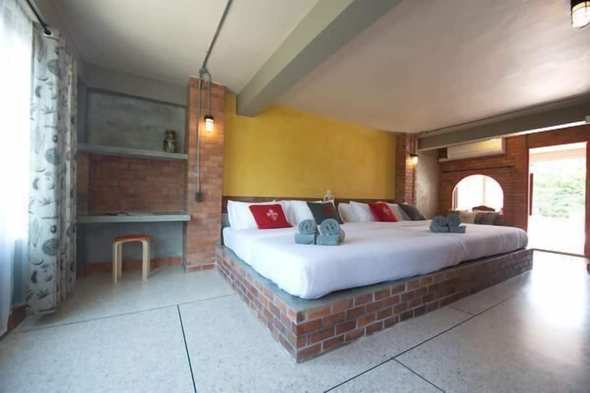 Quarto com cama de tijolo, roupa de cama branca e um banquinho pequeno.