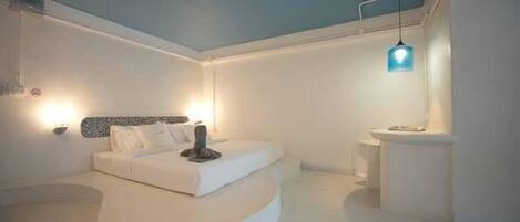 Quarto moderno com cama curva, um espaço para sentar e banheiro com pia e espelho brancos.