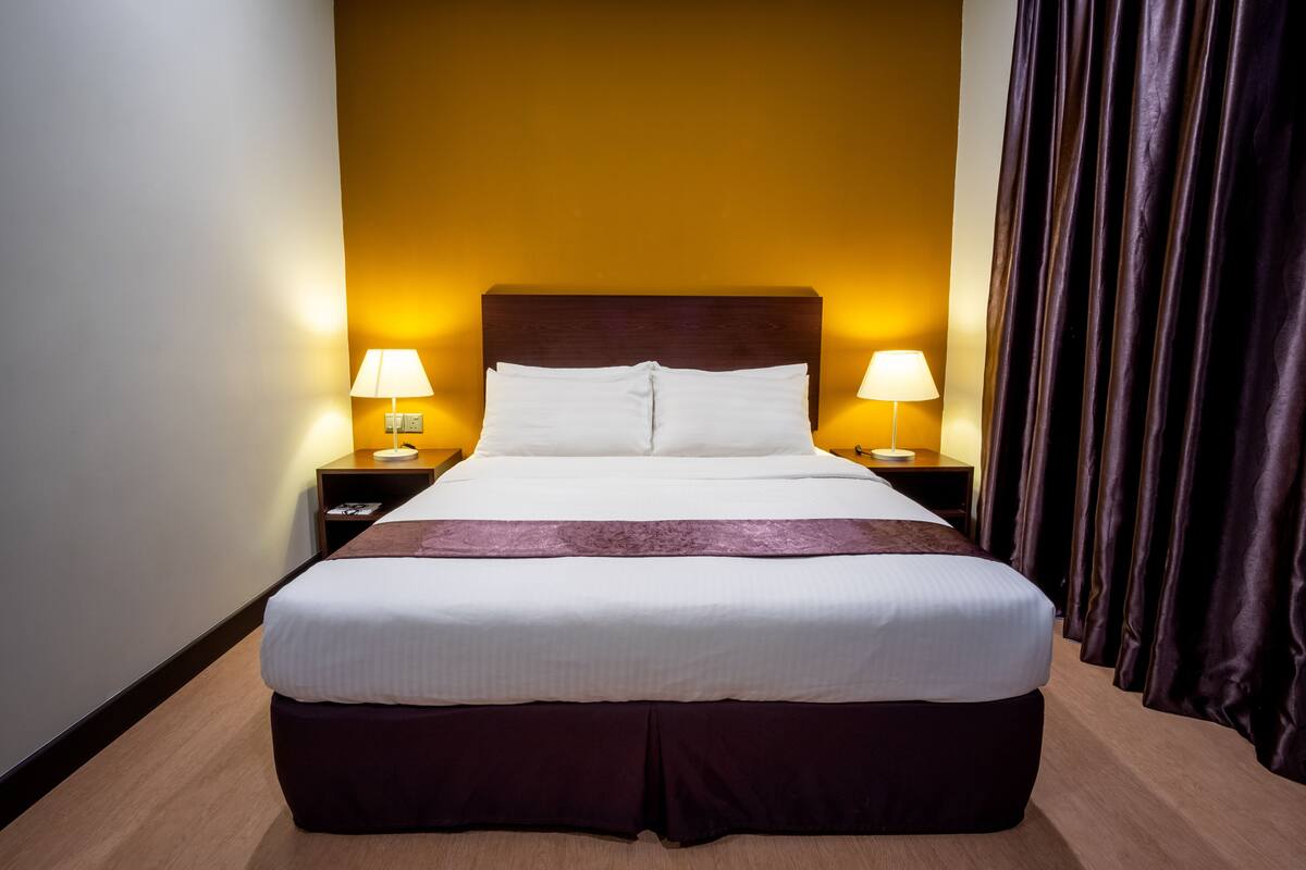 A hotel room with a large bed, two bedside lamps, a dark headboard, and a curtain on the right.