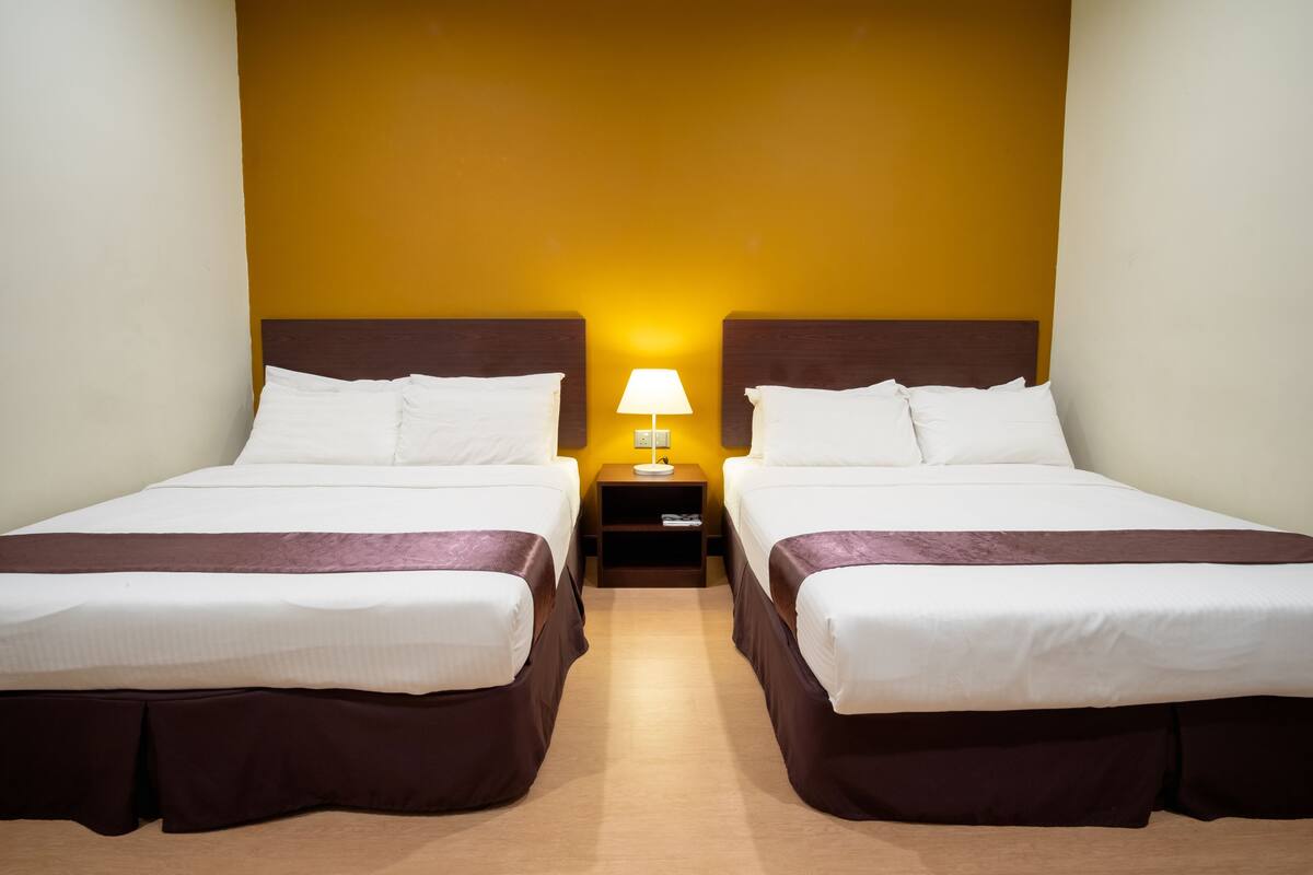 A hotel room with two beds, a nightstand with a lamp, and a yellow accent wall.