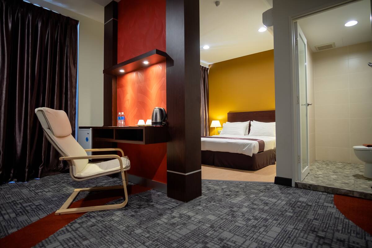 A hotel room with a bed, a chair, a desk with a lamp, a shower area, and a bathroom.