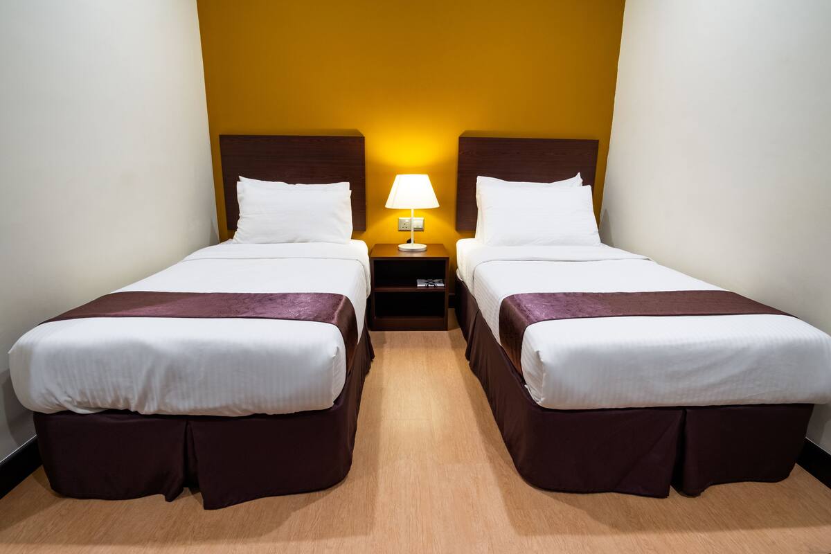 A hotel room with two single beds, a nightstand with a lamp, and a wooden floor.