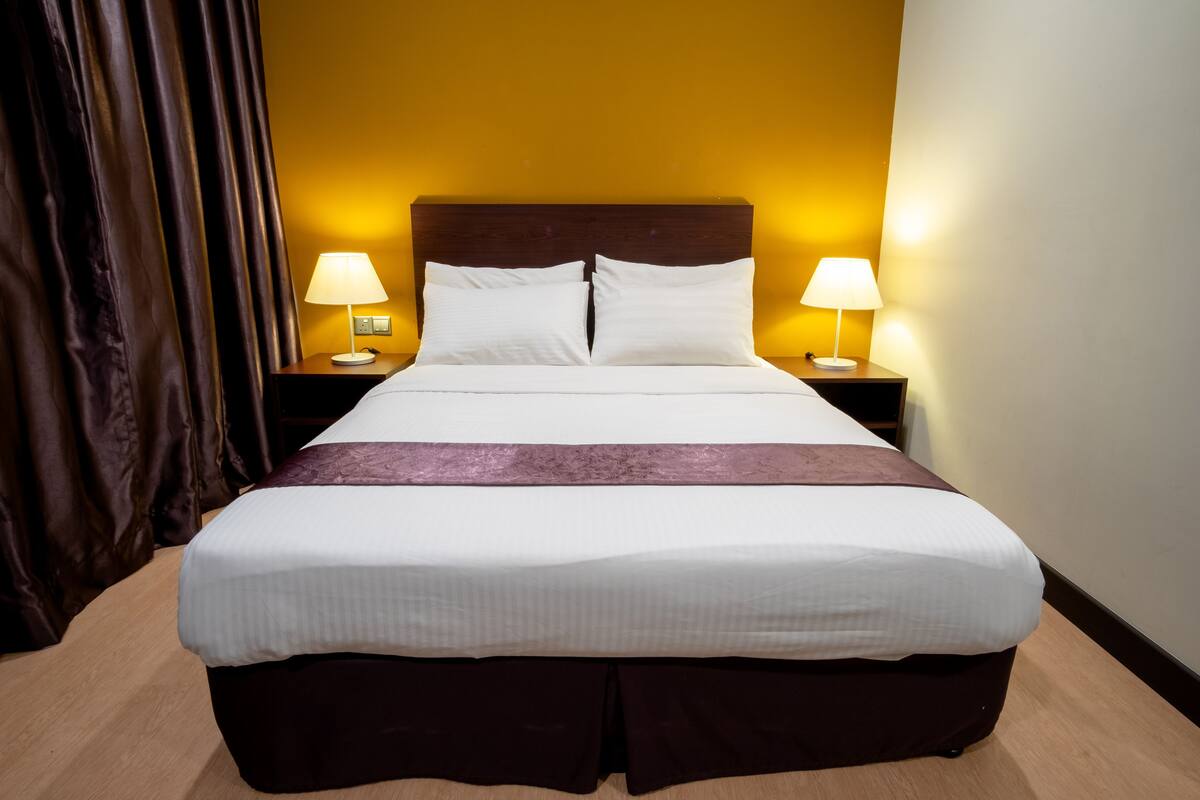 A neatly made bed with white linens and a brown skirt, two bedside lamps, and a dark curtain.