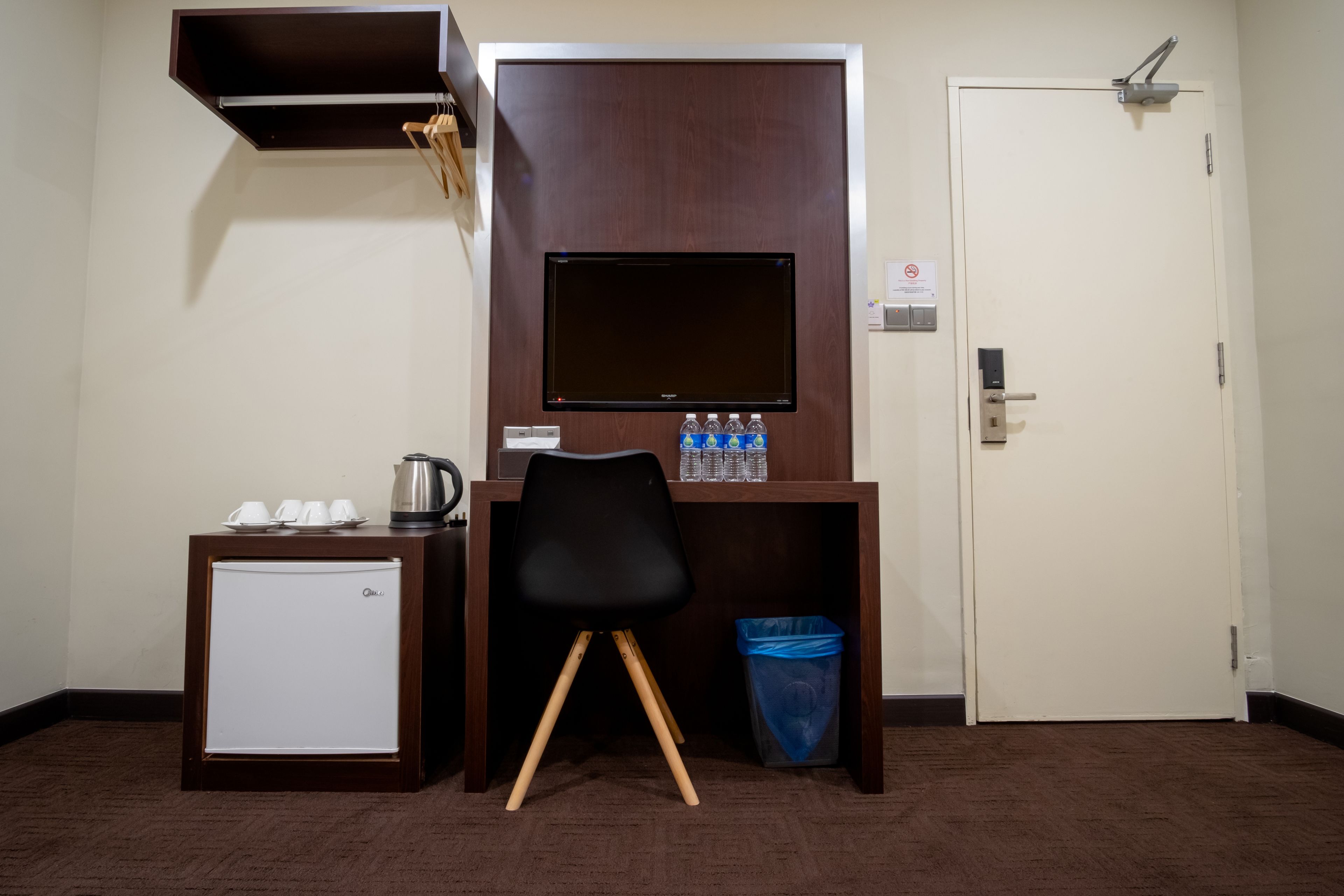Una habitación de hotel pequeña con un refrigerador, un escritorio con monitor de computadora, una silla y un cesto de basura.