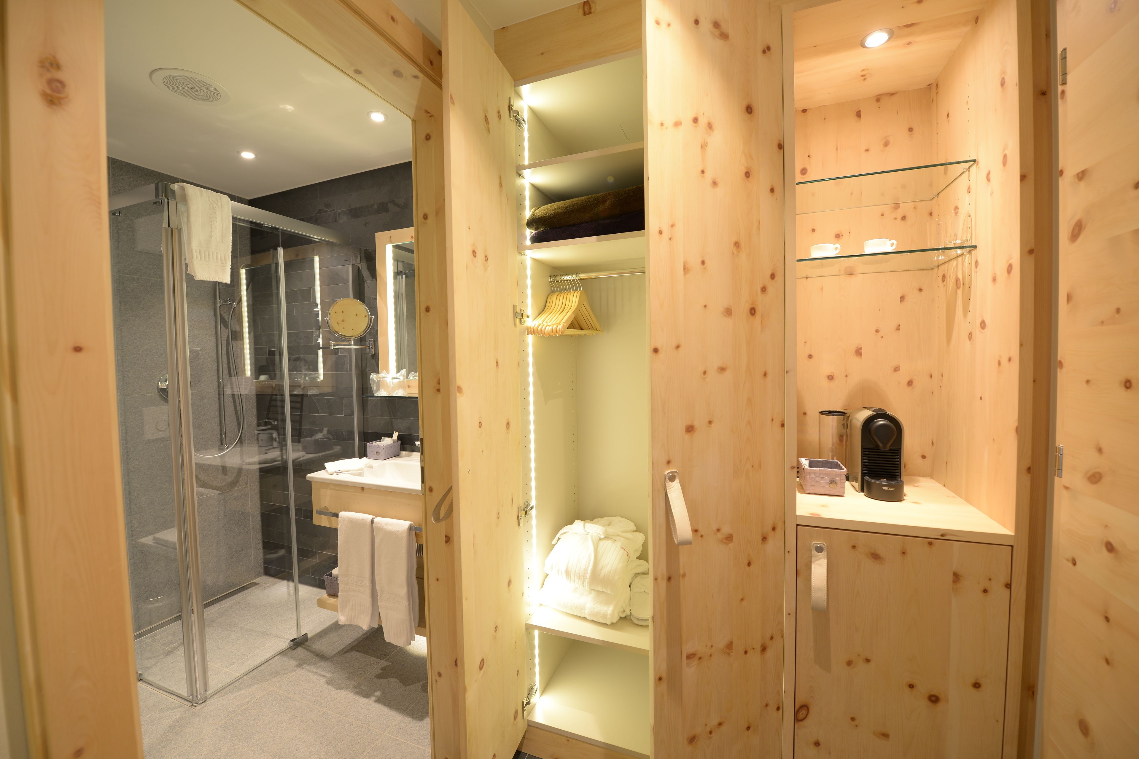 Ein Badezimmer aus Holz mit einer Duschkabine aus Glas, einem Waschtisch mit Spiegel sowie Regalen mit Handtüchern und Pflegeprodukten.