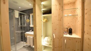 Ein Badezimmer aus Holz mit einer Duschkabine aus Glas, einem Waschtisch mit Spiegel sowie Regalen mit Handtüchern und Pflegeprodukten.