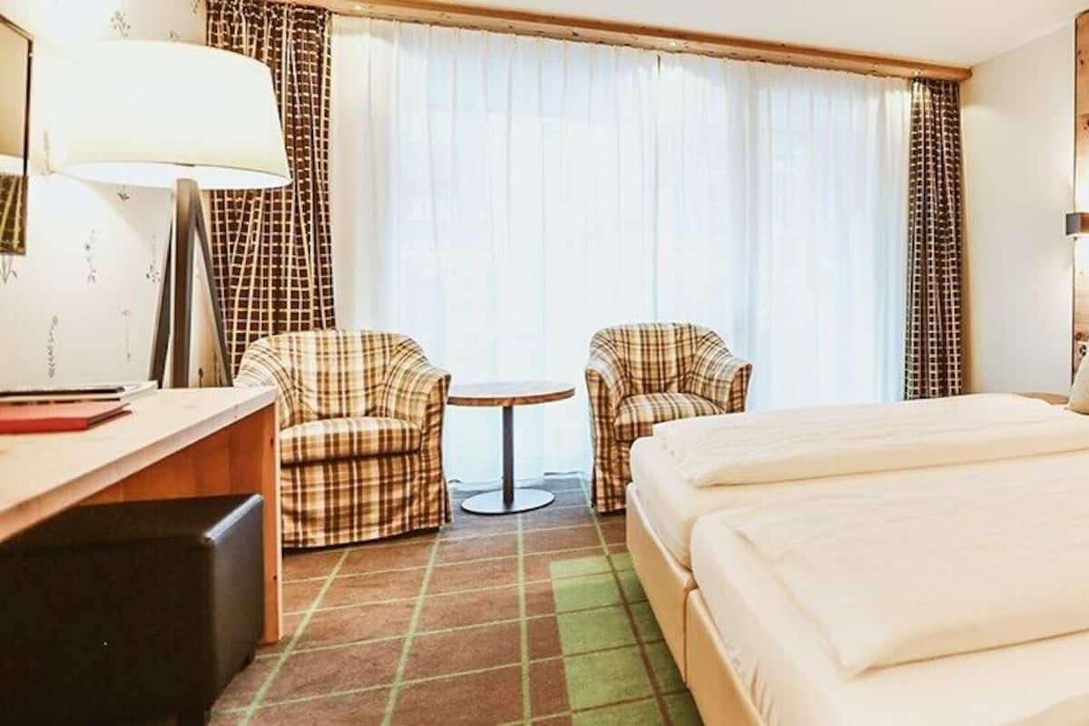 A hotel room with two beds, a desk with a lamp, two plaid chairs, and a small table.