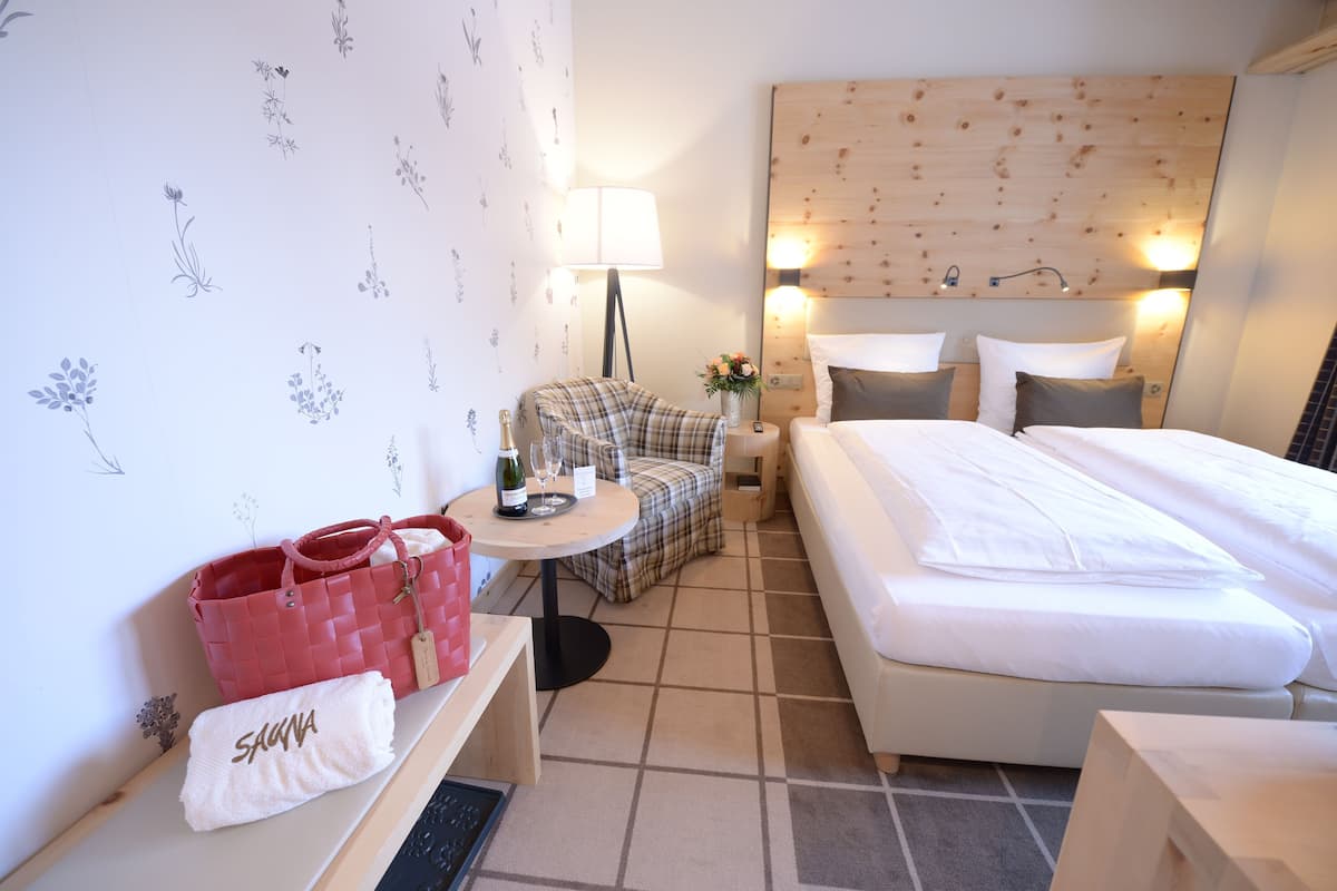 A hotel room with a bed, a chair, a small table with a bottle and glasses, a red bag, and a white towel.