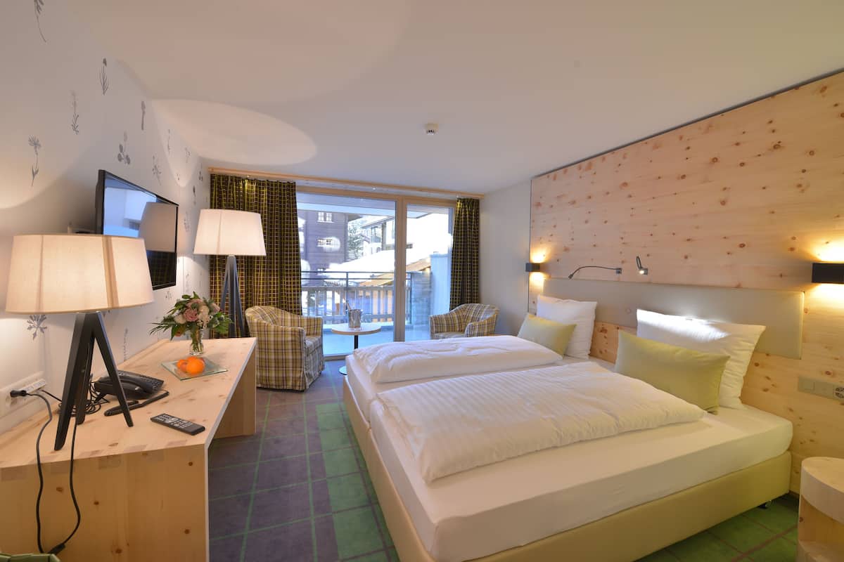 A hotel room with a bed, a desk with a computer and phone, a chair, and a view of the outside.