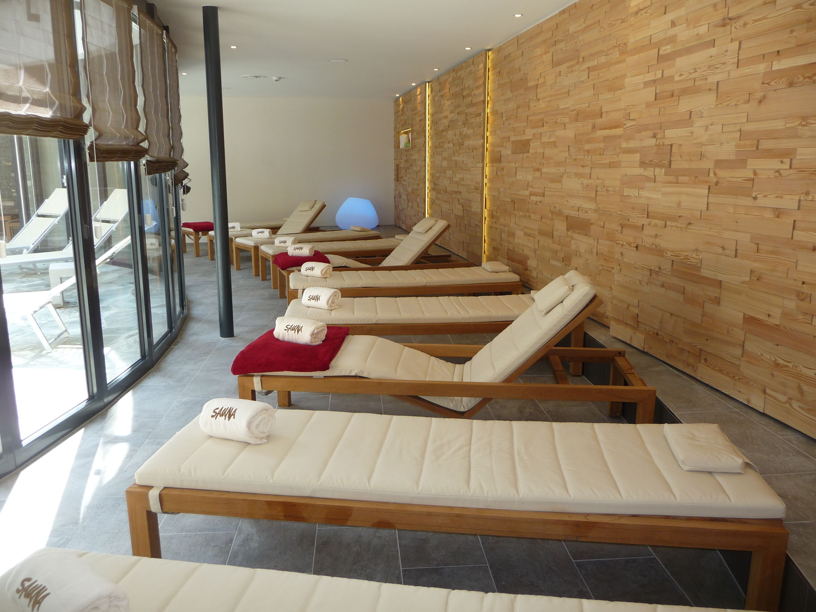 A row of wooden sun loungers with towels, a stone wall, and a large window.