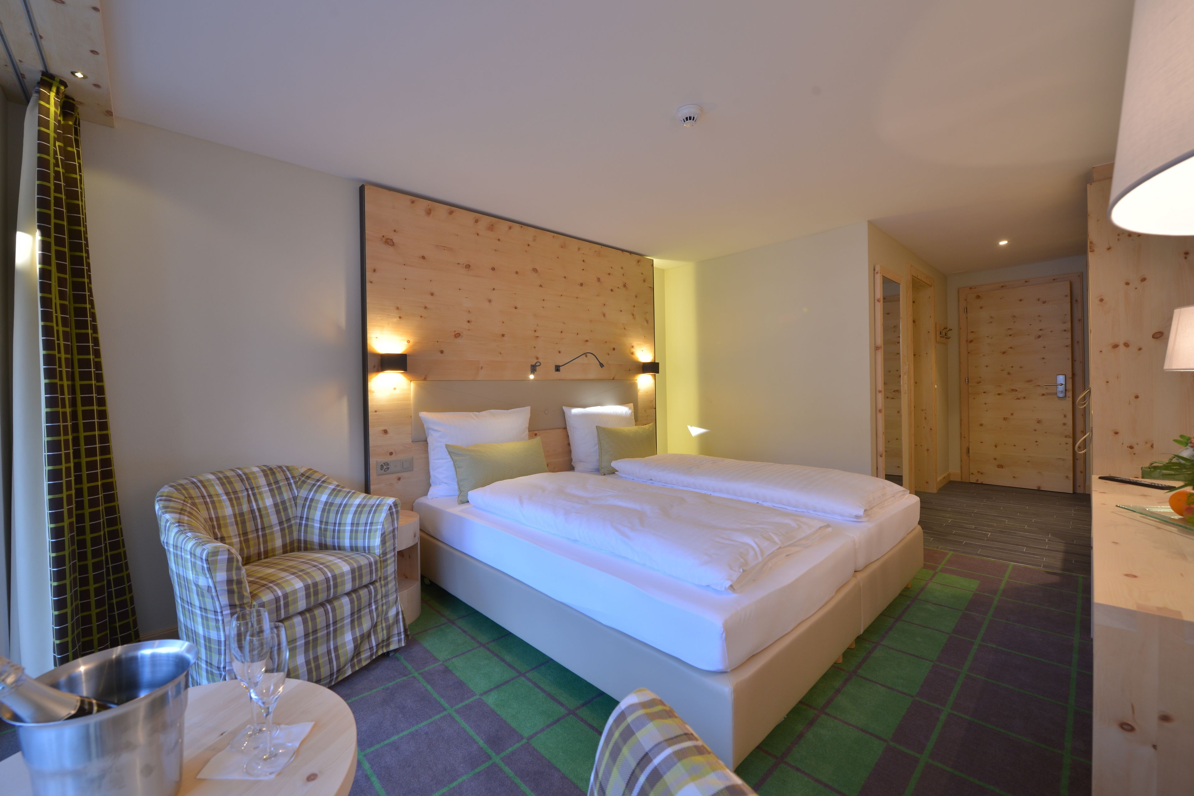 A hotel room with a large bed, a chair, a small table with a glass, and a wooden headboard.