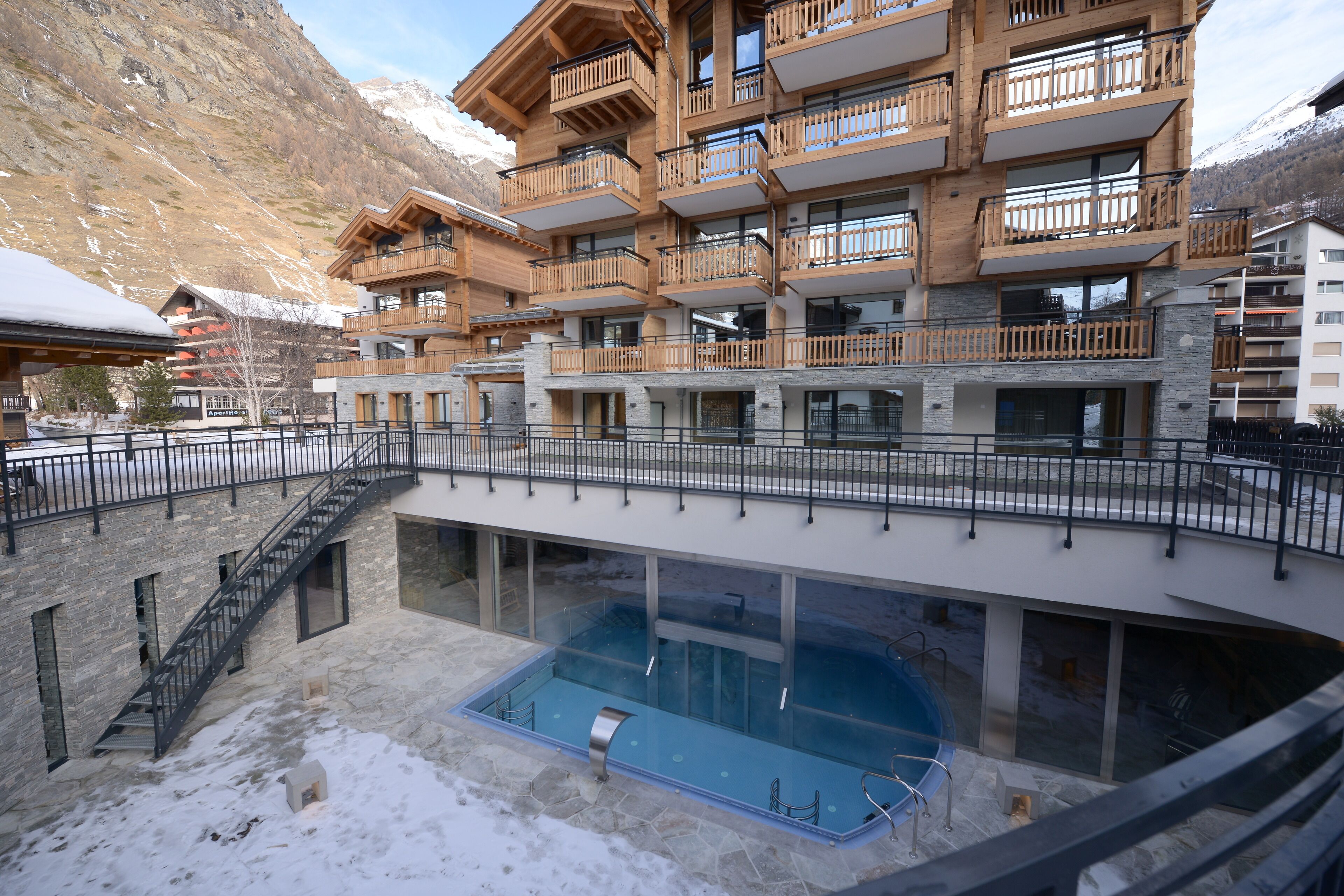 A multi-story hotel with a pool area, wooden balconies, and a mountainous backdrop.