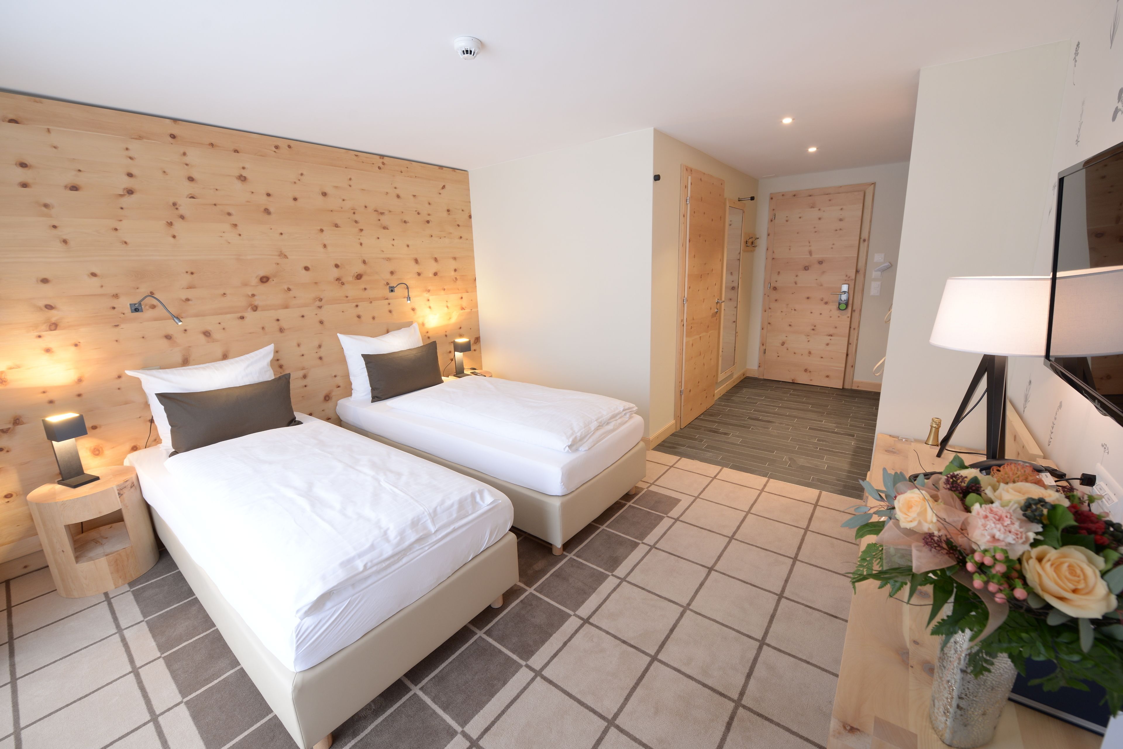 Una camera d'albergo con due letti singoli, una testiera di legno, lampade ai comodini, una TV a schermo piatto, un vaso con fiori e un pavimento piastrellato.