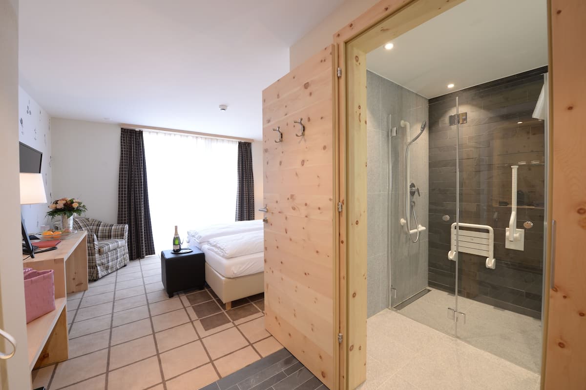 A modern hotel room with a bed, a desk, a small table with a bottle, a chair, a sofa, and a shower area with glass doors.
